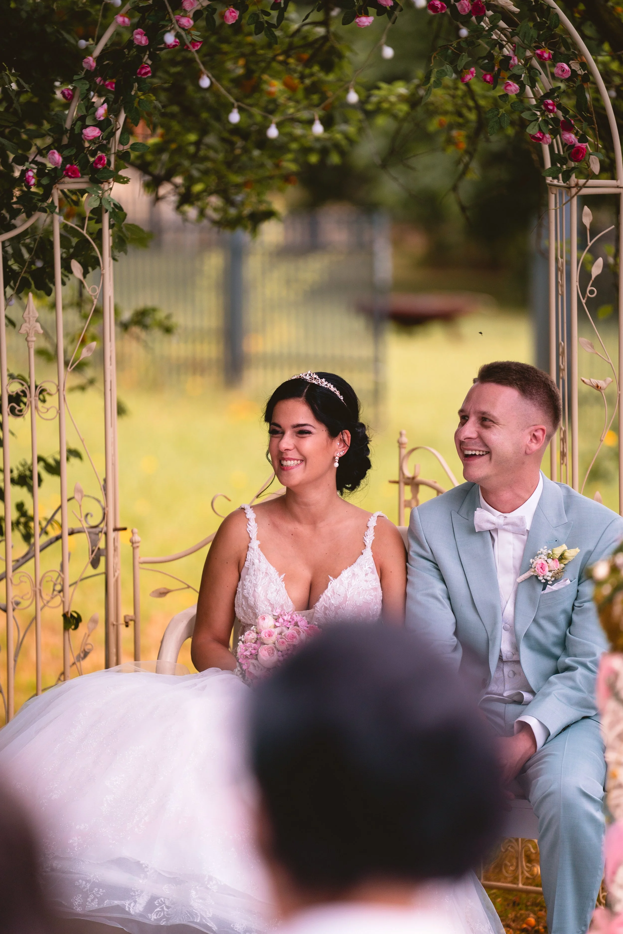 Ein Hochzeitsfoto eines glücklichen Paares, sitzend im Garten unter einer Blumenbogen-Hochzeitspforte, umgeben von pinken Rosen und grünen Blättern.