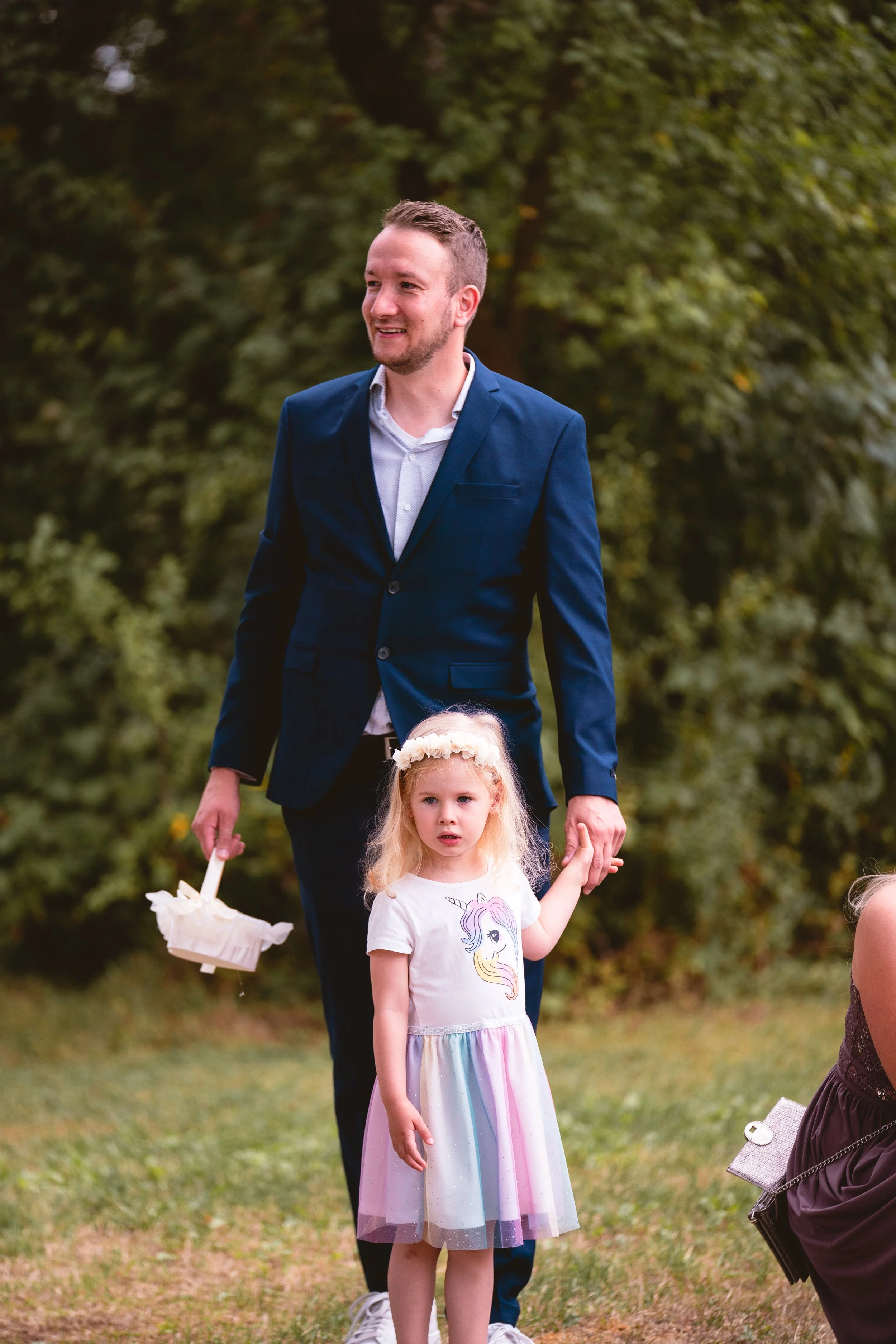 Ein Mann in einem blauen Anzug hält die Hand eines kleinen Mädchens mit Regenbogen- und Einhorn-Print-Kleid, während sie im Freien stehen.