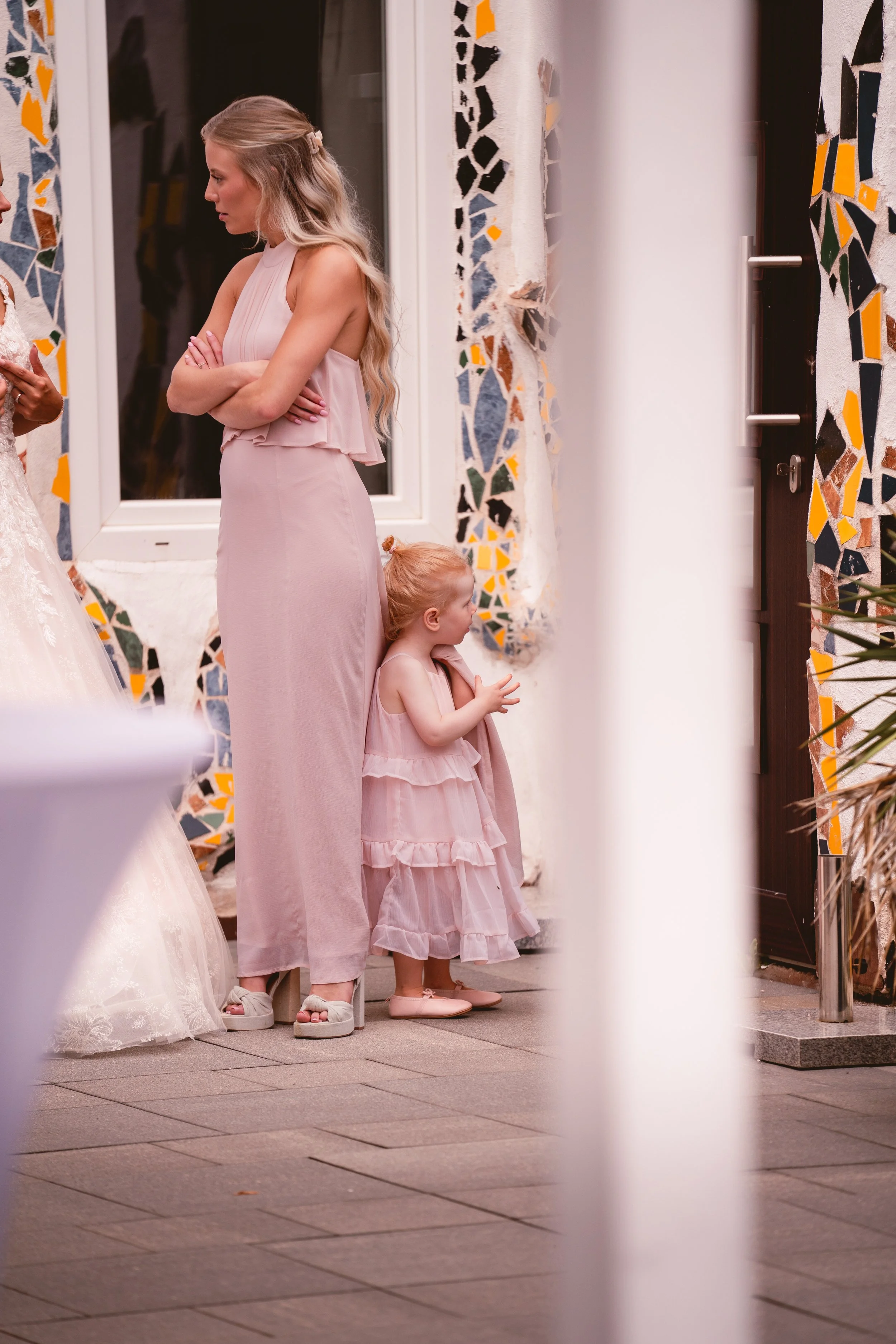 Zwei junge Mädchen in eleganten Kleidern, eines in rosa, stehen vor einer farbenfrohen Wand aus Mosaiksteinen. Sie sind im Gespräch, während sie in einem Eingangsbereich stehen, flankiert von einem weiß gehaltenen Rahmen mit einer Tür im Hintergrund.
