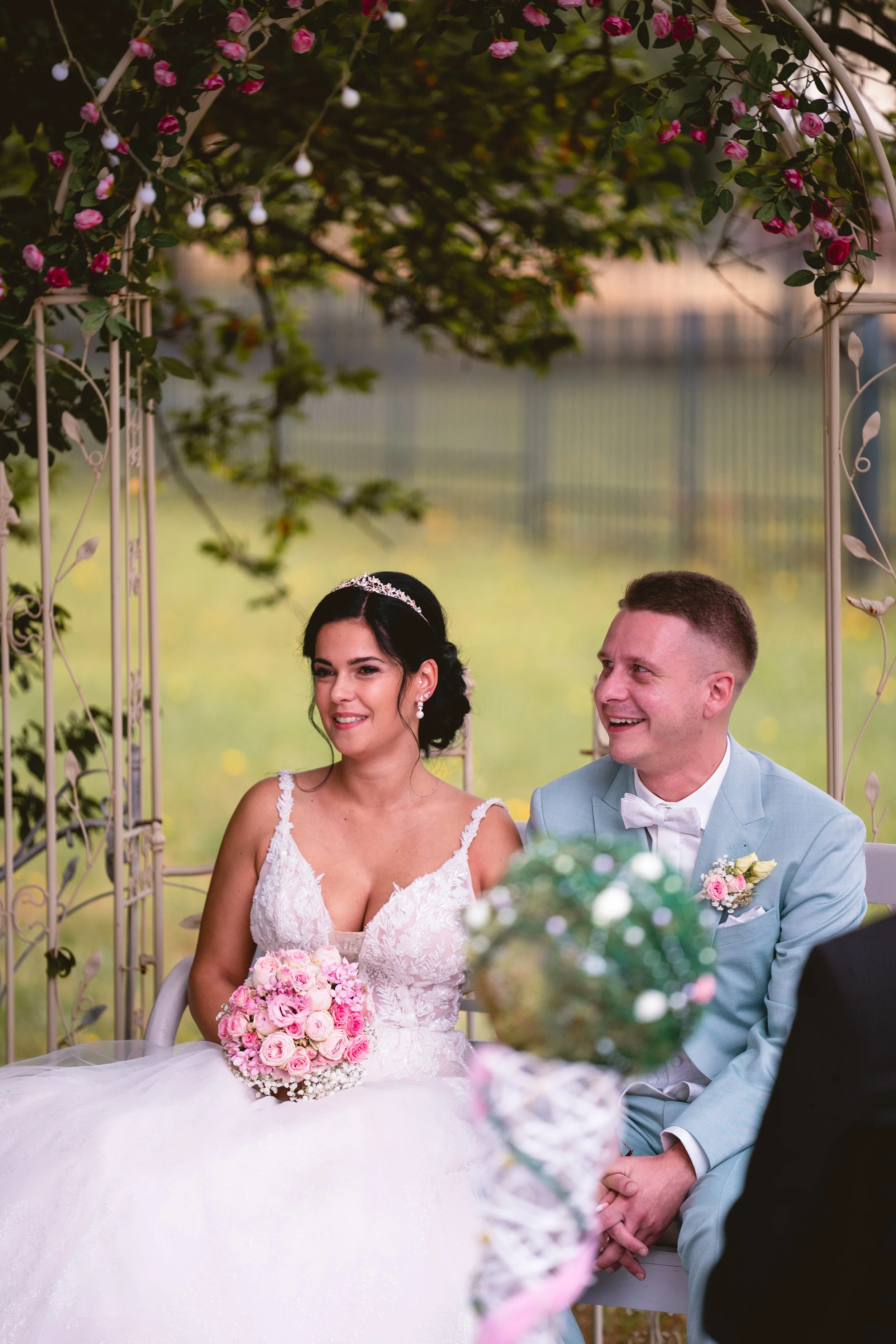 Ein Brautpaar bei einer Hochzeit, sitzend unter einem blumengeschmückten Bogen, die Braut hält einen Blumengebinde und trägt ein weißes Hochzeitskleid, der Bräutigam trägt einen hellblauen Anzug. Im Hintergrund sind Bäume und eine grüne Wiese sichtba