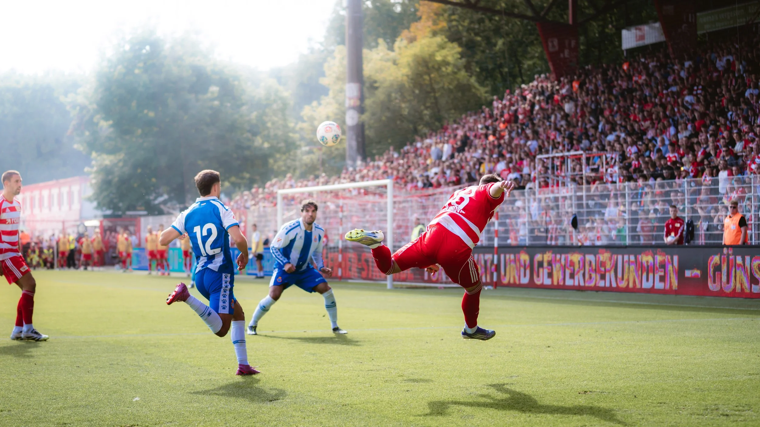 Fußballspiel mit Spielern in blauen und roten Trikots, Torwart springt, im Hintergrund Jubelnde Fans im Stadion.