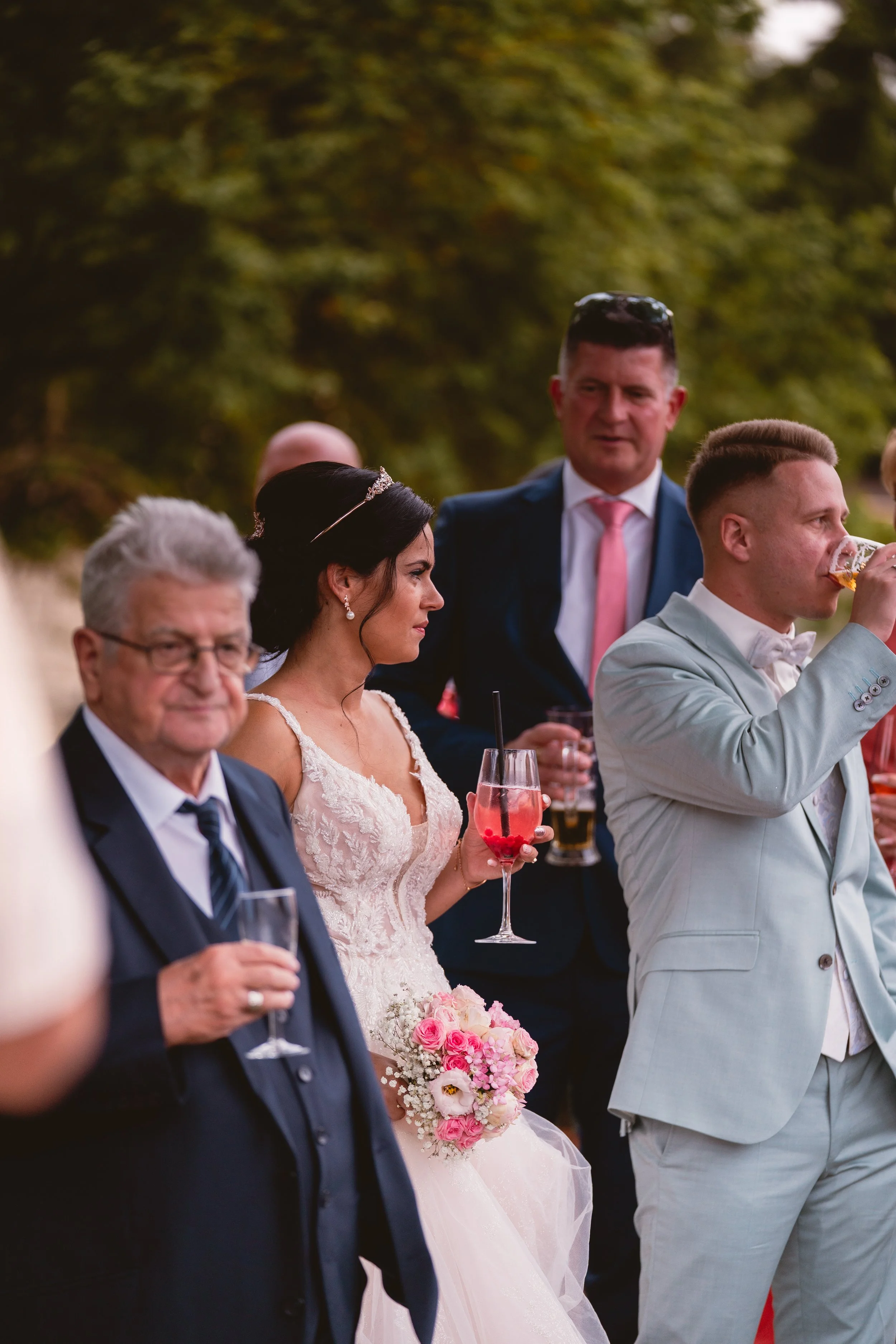 Menschen bei einer Hochzeitfeier, die im Freien stattfinden, mit einem Brautpaar in der Mitte, einer Frau in Brautkleid mit Blumenstrauß und Männern in Anzügen, einige trinken Getränke.