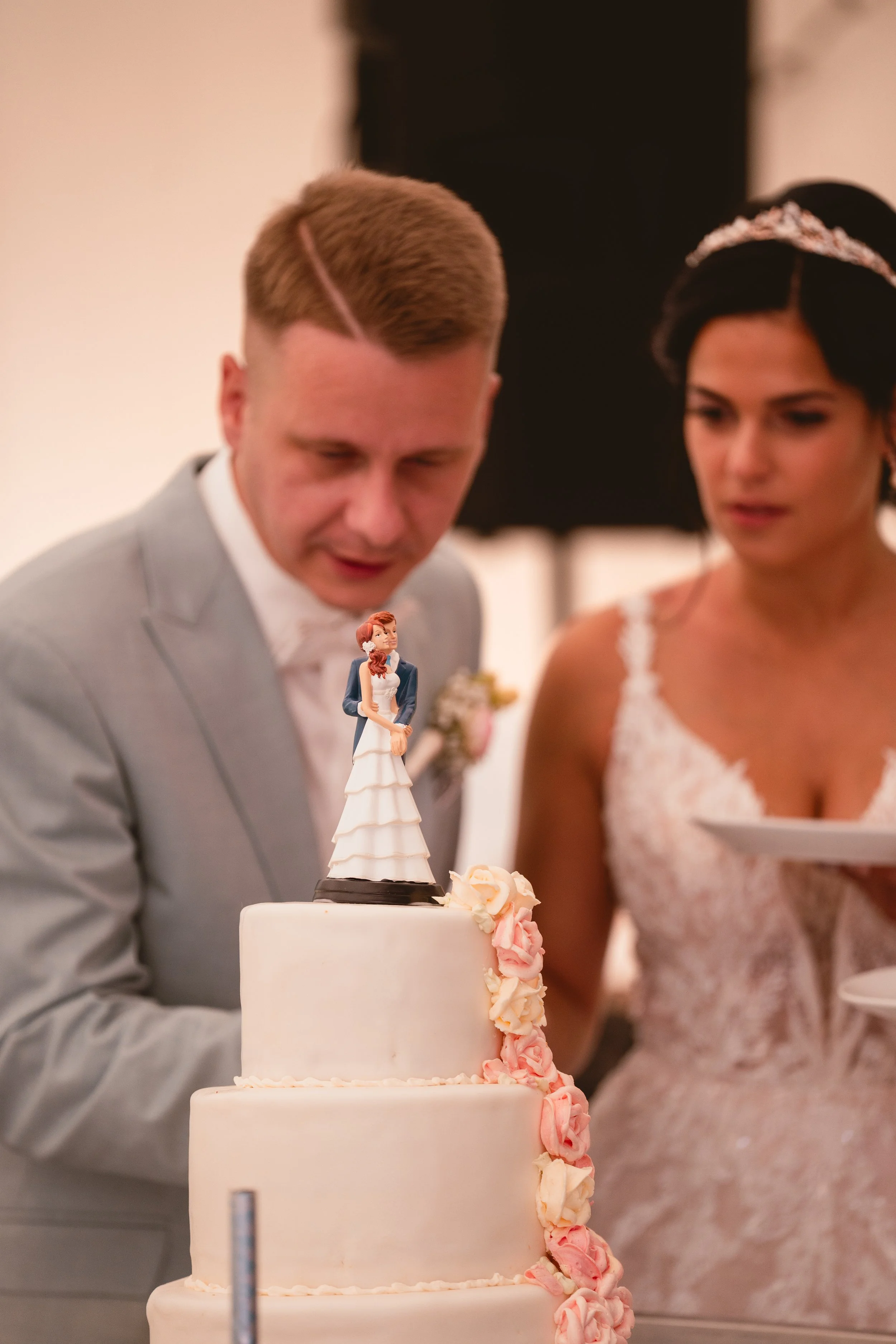 Ein Brautpaar bei einer Hochzeitstorte, der Bräutigam trägt einen hellgrauen Anzug und die Braut trägt ein weißes Spitzenkleid und eine Tiara, die Torte ist mehrstöckig mit weißen und rosa Dekorationen.