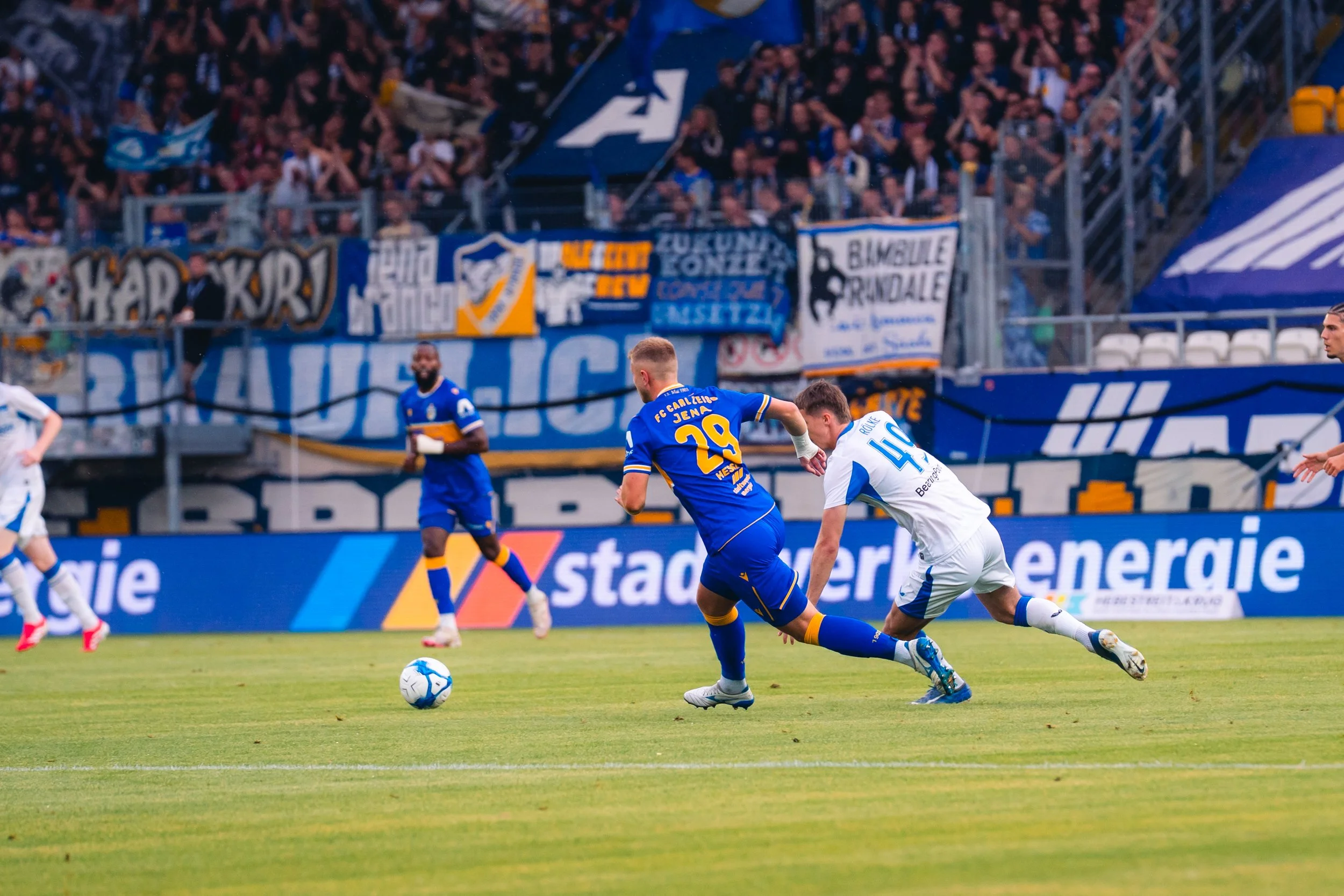 Fußballspieler in blauer und weißer Trikot während eines Spiels auf dem Spielfeld, im Hintergrund ein ausverkauftes Stadion mit Fans und Bannern.