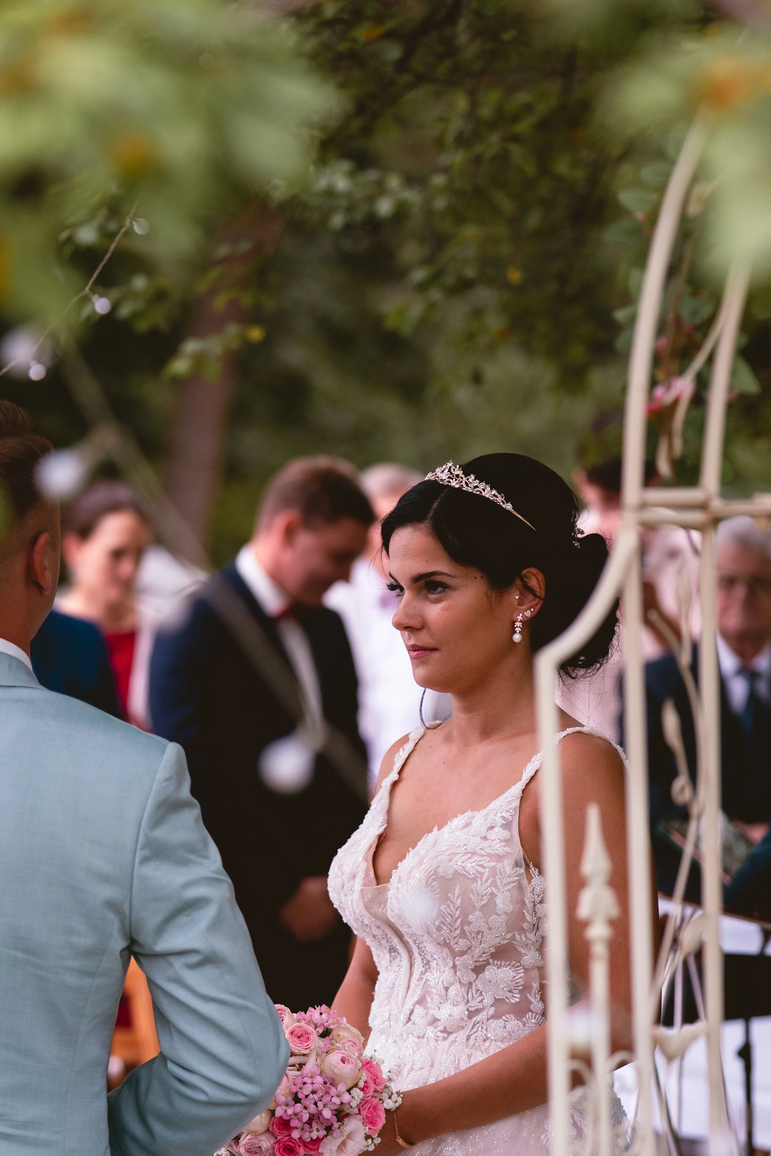 Eine Braut und ein Bräutigam bei einer Hochzeitszeremonie im Freien, der Fokus liegt auf der Braut, die ein weißes Hochzeitskleid trägt und eine Tiara trägt, während sie mit dem Bräutigam spricht, umgeben von Gästen und Dekorationen.