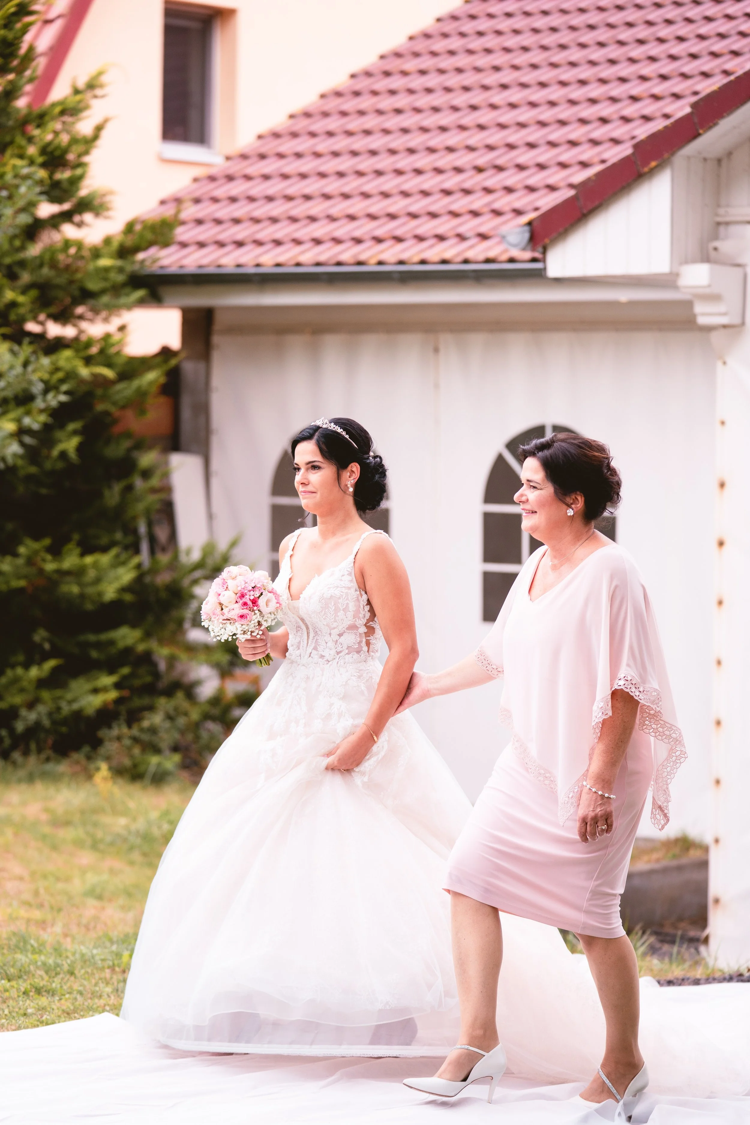 Eine braut in einem weißen Kleid mit Blumenstrauß, begleitet von einer Frau in hellrosa Stoff, verlässt das Haus nach der Hochzeit.