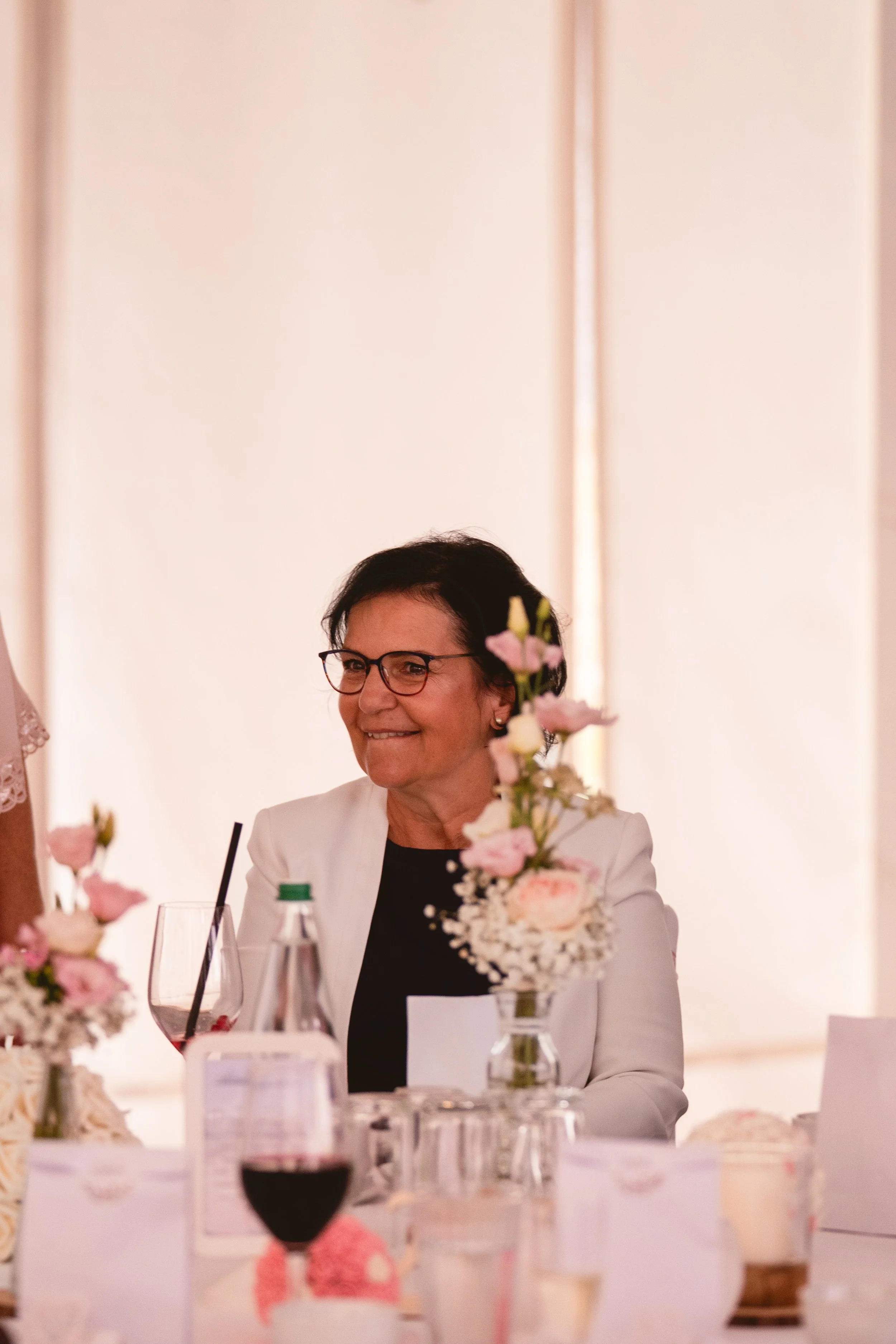 Eine Frau mit dunklem Haar und Brille sitzt an einem gedeckten Tisch bei einem festlichen Anlass, umgeben von Blumen und Wein. Sie trägt einen weißen Blazer und hat ein freundliches Lächeln.