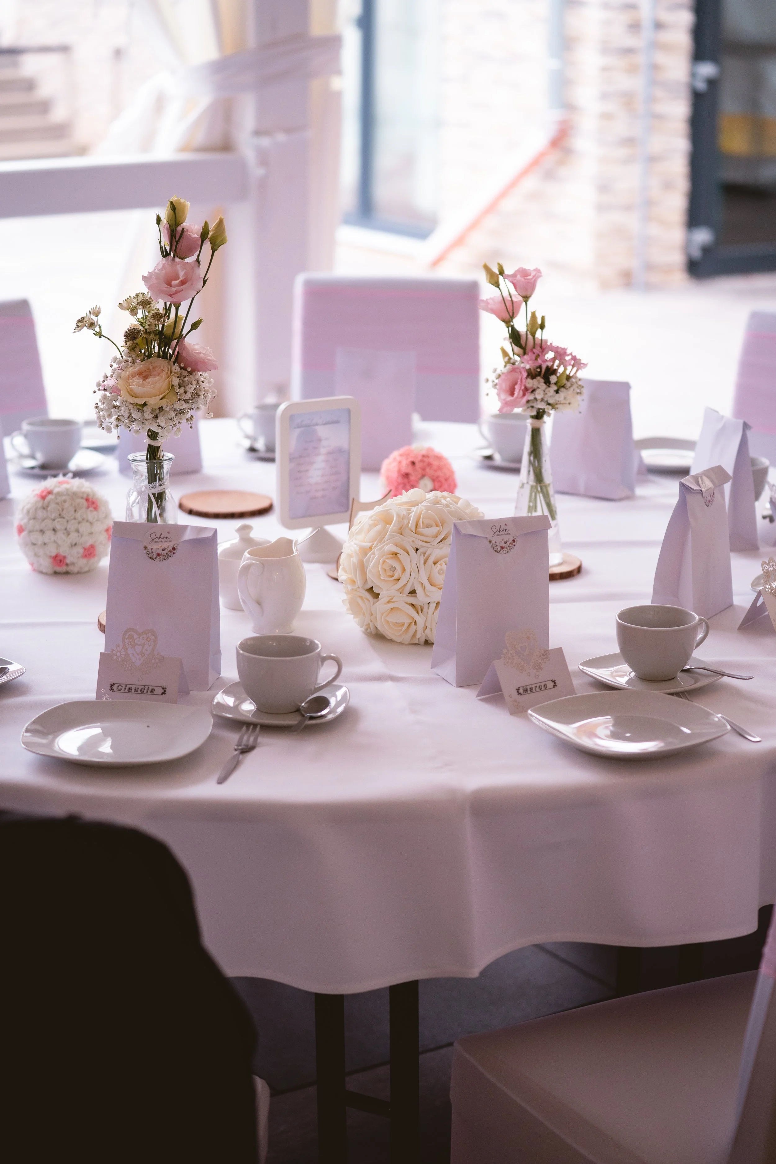Tisch mit weißer Tischdecke, Kerzen, Blumenarrangements in Gläsern, Tassen, Tellern und Platzkarten, vermutlich für eine Hochzeit oder Feierlichkeit, im Hintergrund eine große Fensterscheibe mit Blick nach draußen