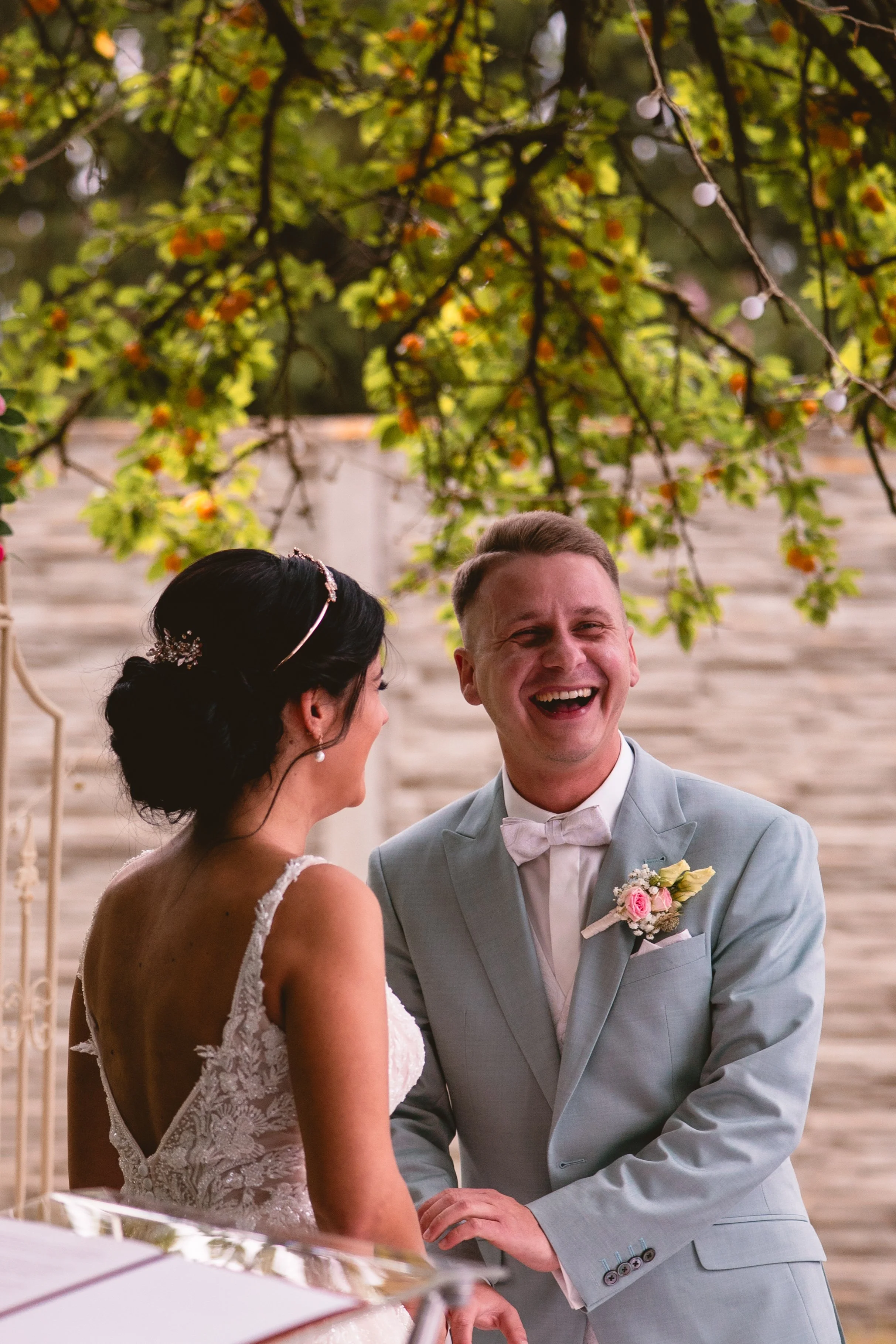 Ein glückliches Brautpaar bei ihrer Heirat, der Bräutigam trägt einen hellgrauen Anzug mit rosa Blumen im Revers, die Braut trägt ein elegantes Spitzenkleid. Sie stehen unter einem Baum mit grünen Blättern und kleinen orangen Beeren, im Hintergrund e