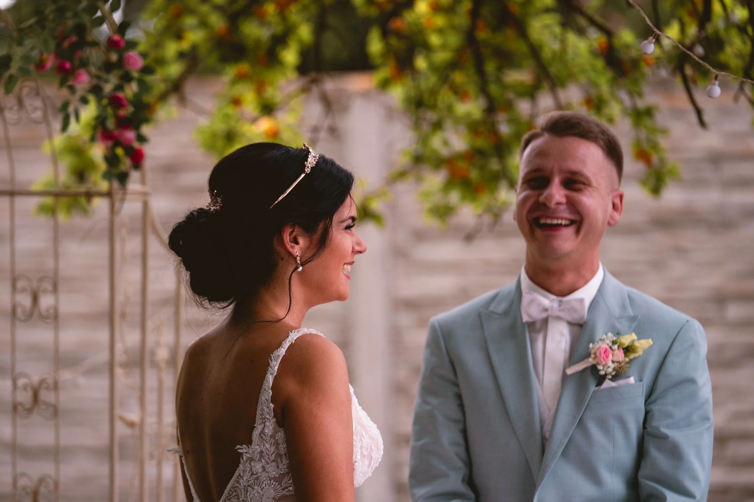 Ein Brautpaar bei ihrer Hochzeit, lachend und sich gegenseitig zugewandt, hinter ihnen hängen grüne Blätter und Girlanden, eine festliche Atmosphäre.