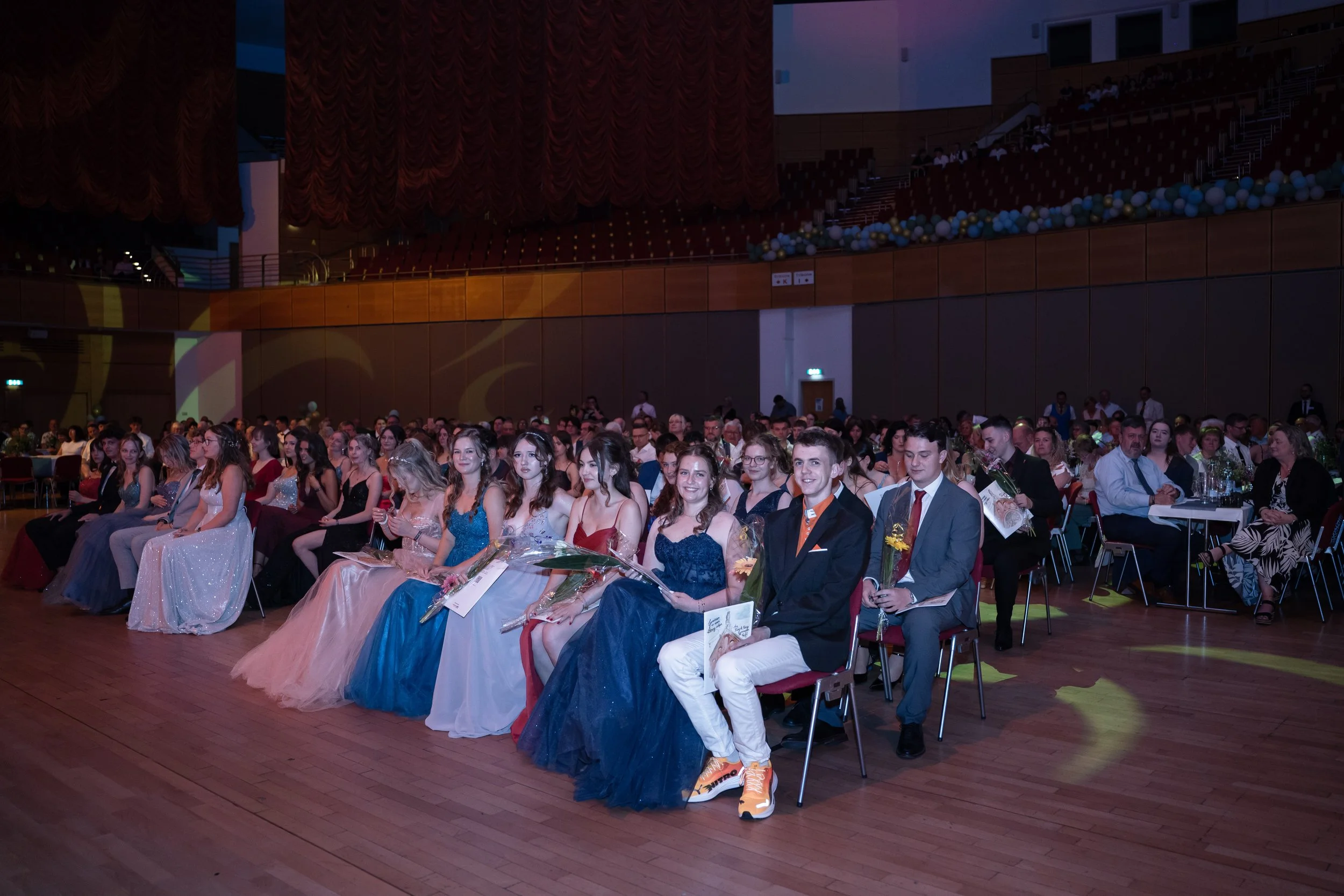 Junge Menschen bei einer Abschlussfeier oder Gala sitzen in einem Veranstaltungssaal, viele tragen formelle Kleidung, einige halten Blumen und Zertifikate in den Händen.