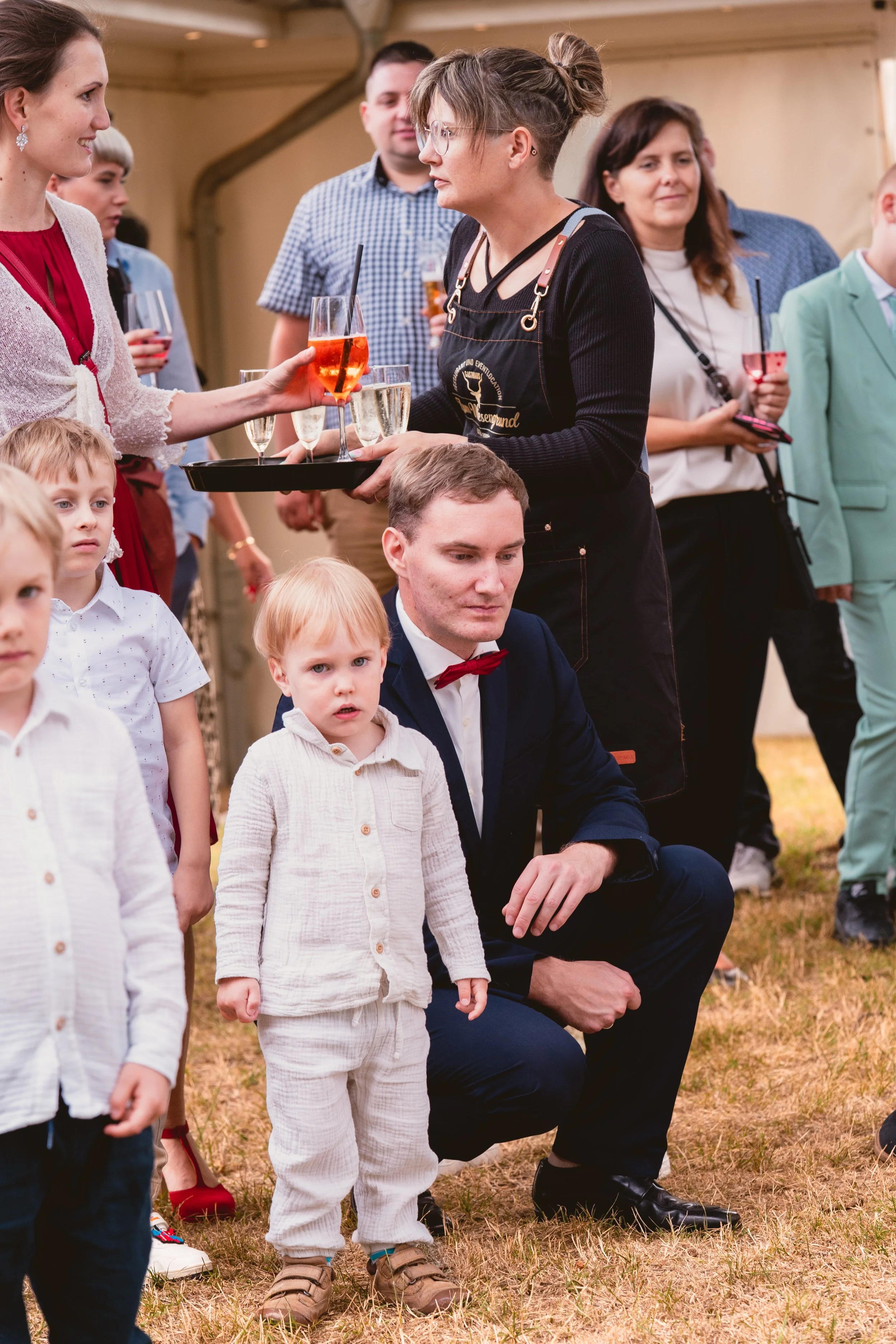 Menschen bei einer Veranstaltung, Mädchen serviert Getränke, Kinder und Erwachsene im Hintergrund, draußen auf Gras, vermutlich in einem Festzelt.