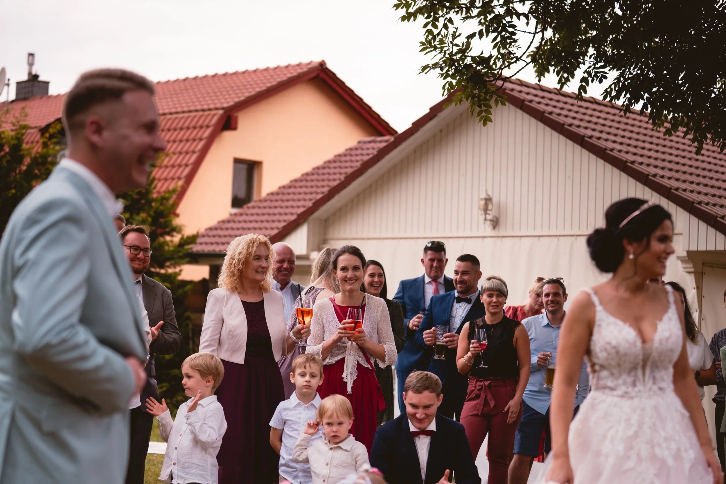 Hochzeitsparty im Freien mit lachenden Erwachsenen und Kindern, im Vordergrund eine Braut in weißem Kleid, im Hintergrund ein Haus mit roten Ziegeln. Es ist ein sonniger Tag.