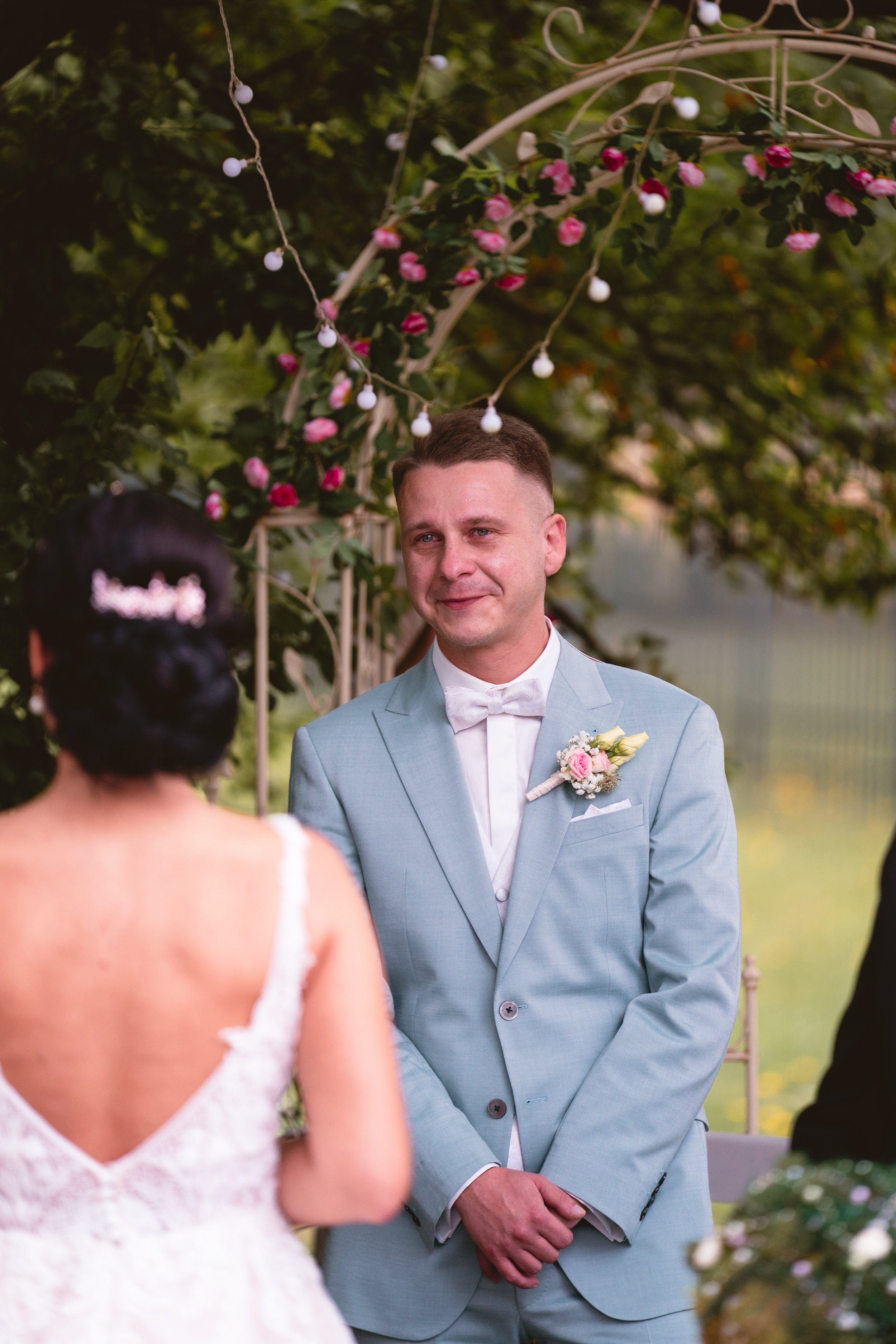 Ein Mann in einem hellblauen Anzug bei einer Hochzeitszeremonie, im Hintergrund blühende Blumen und eine Girlande. Er sieht glücklich und emotional aus.