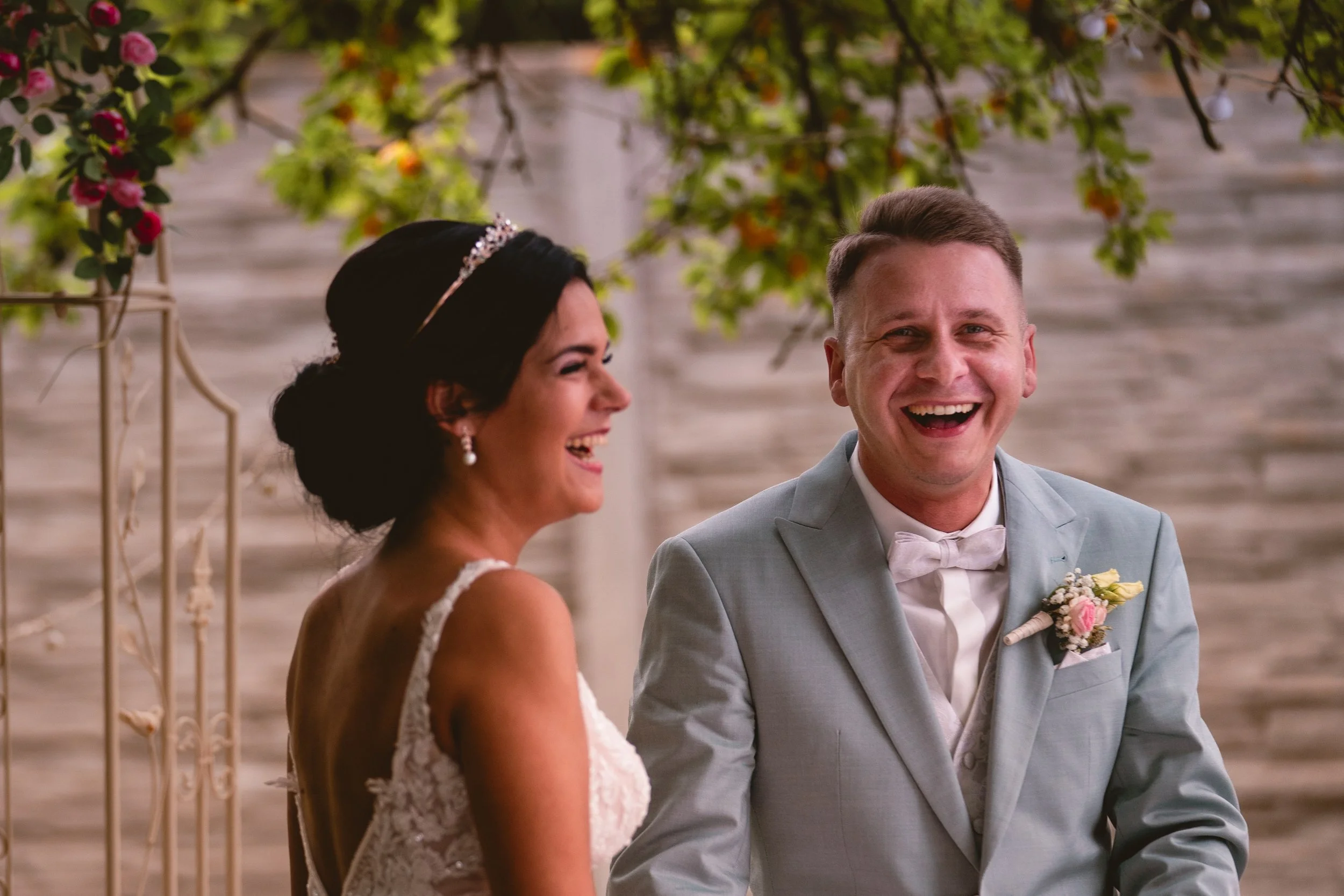 Ein lachendes Paar bei einer Hochzeit, mit einem Blumenstrauß, im Freien vor einer steinernen Mauer, blühenden Bäumen und dekorativen Blumen.