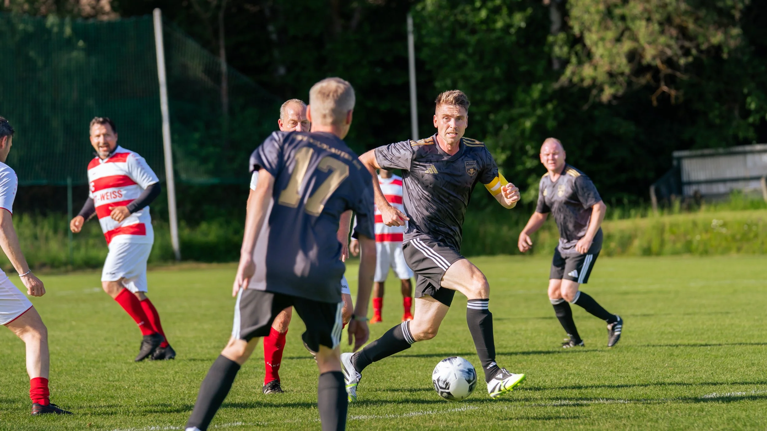 Fußballspiel auf dem Grashof mit mehreren Spielern, ein Mann trägt eine schwarze Uniform mit einem Kapitänsbinde, spielt mit Ball, andere in weißen und roten Trikots sind auf dem Spielfeld.