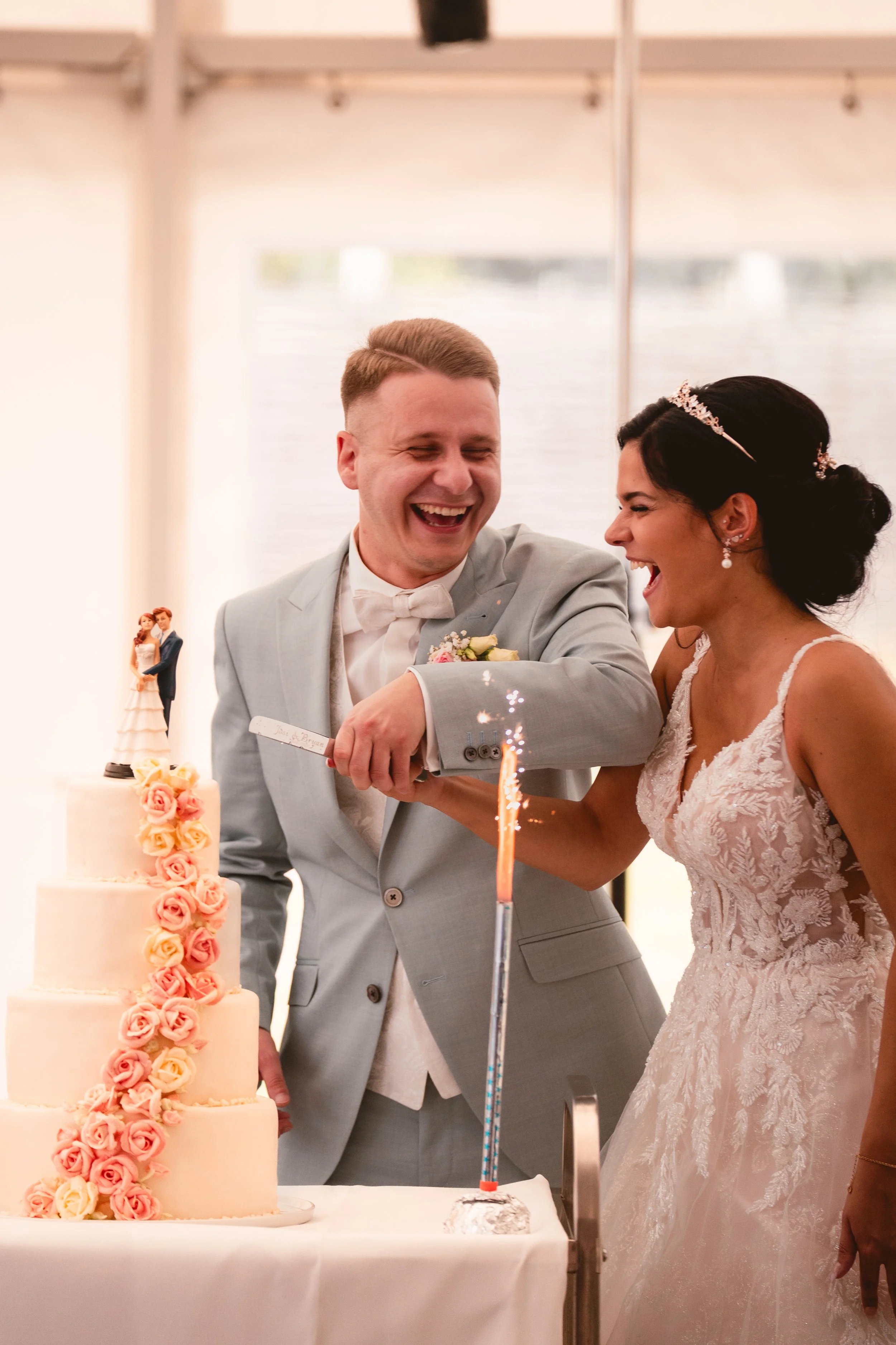 Ein Brautpaar beim Anschneiden der Hochzeitstorte, beide lachen glücklich, die Braut trägt ein weißes Spitzenkleid mit Spaghettiträgern und eine Diadem, der Bräutigam trägt einen hellgrauen Anzug mit Fliege. Im Vordergrund eine mehrstöckige Hochzeits