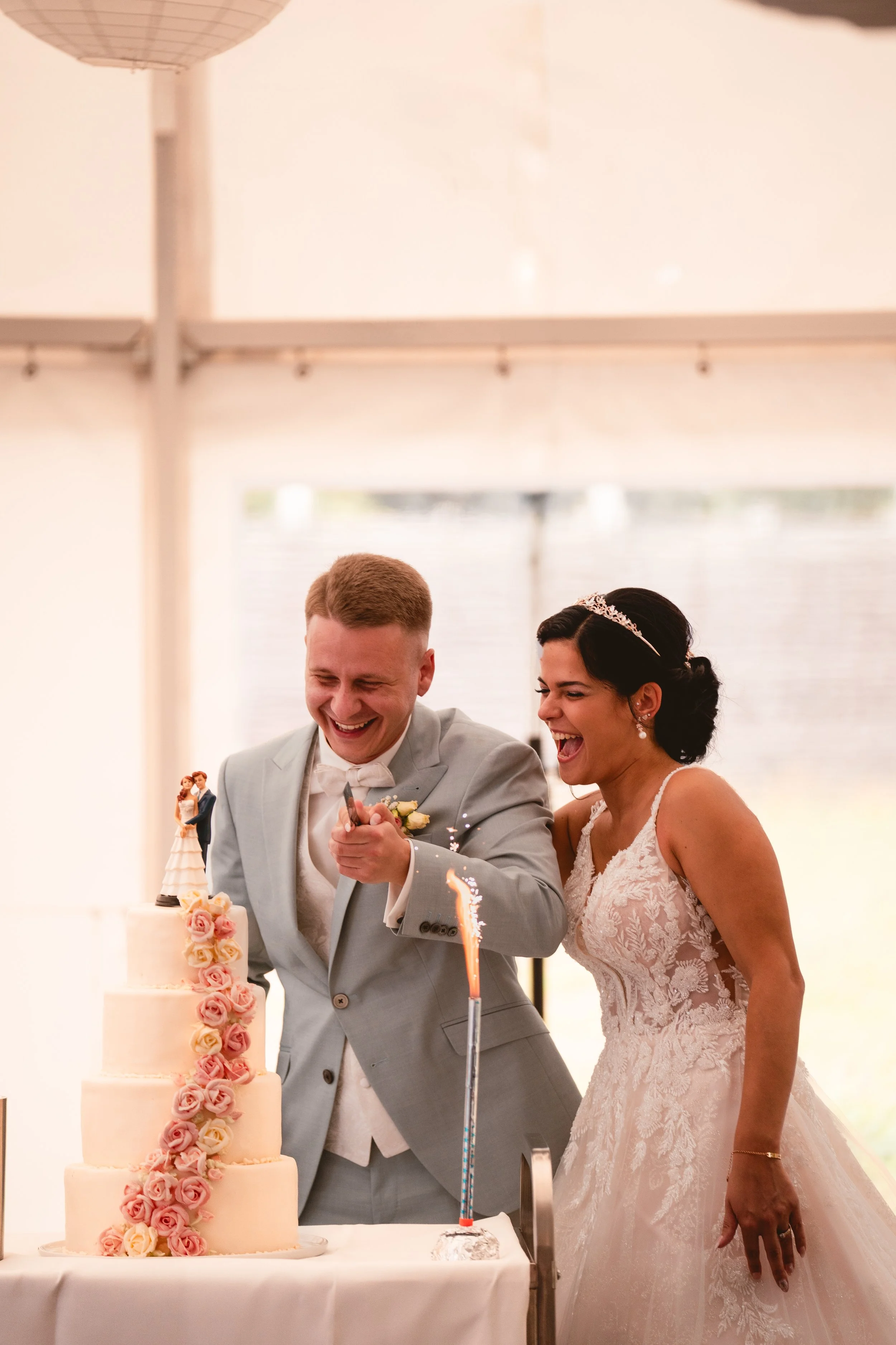 Ein Brautpaar schneidet eine Hochzeitstorte an, beide lachen, im Vordergrund eine mehrstöckige Torte mit rosa und cremefarbenen Blumen. Das Brautpaar trägt elegante Kleidung, der Bräutigam ein hellgrauen Anzug und die Braut ein Spitzenkleid mit Haara