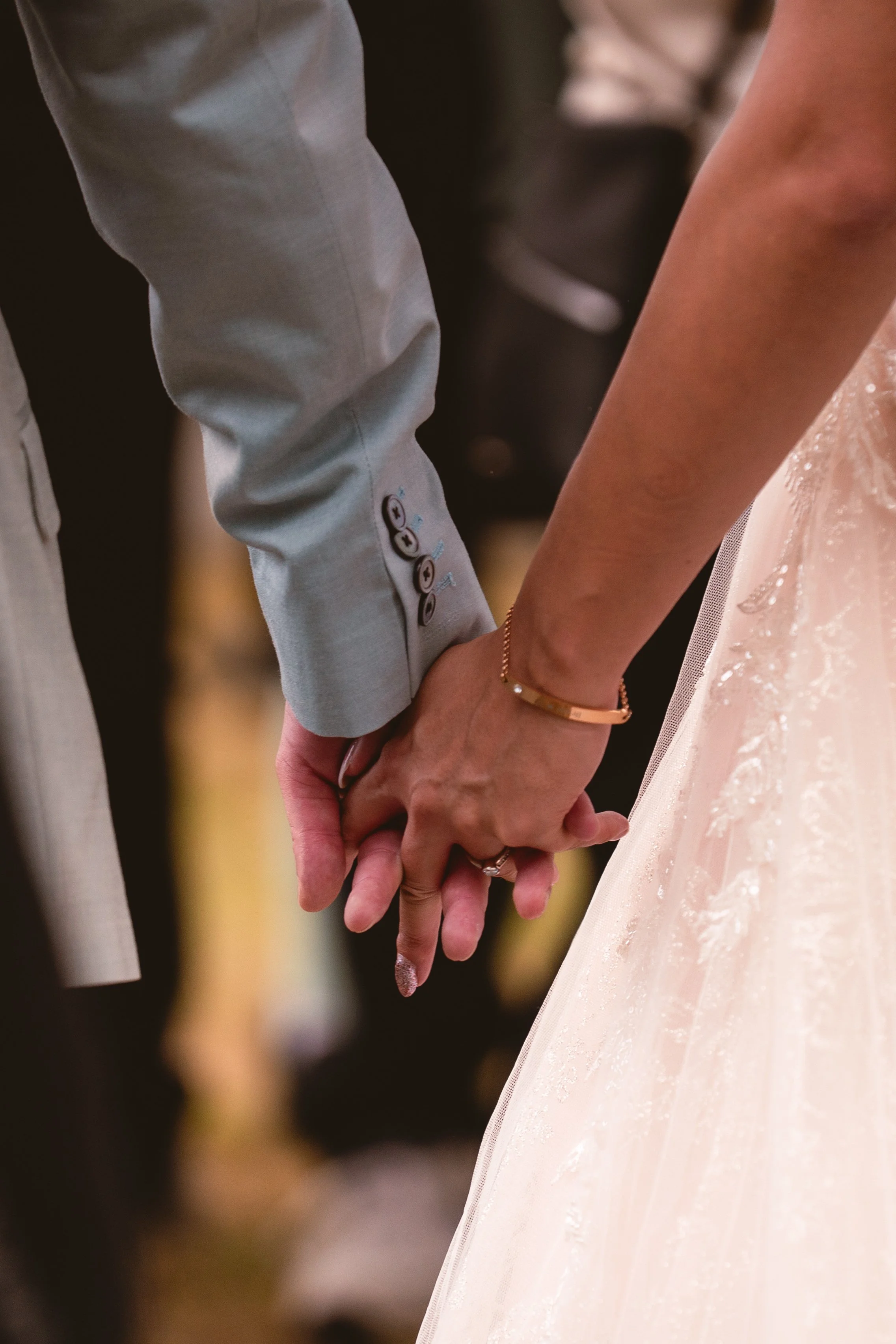 Ein Paar hält sich an den Händen. Die Frau trägt eine goldene Armbanduhr und einen Fingerring. Sie ist in einem pastellfarbenen, verzierten Kleid. Die Hand des Mannes ist zu sehen, er trägt einen hellgrauen Anzug.