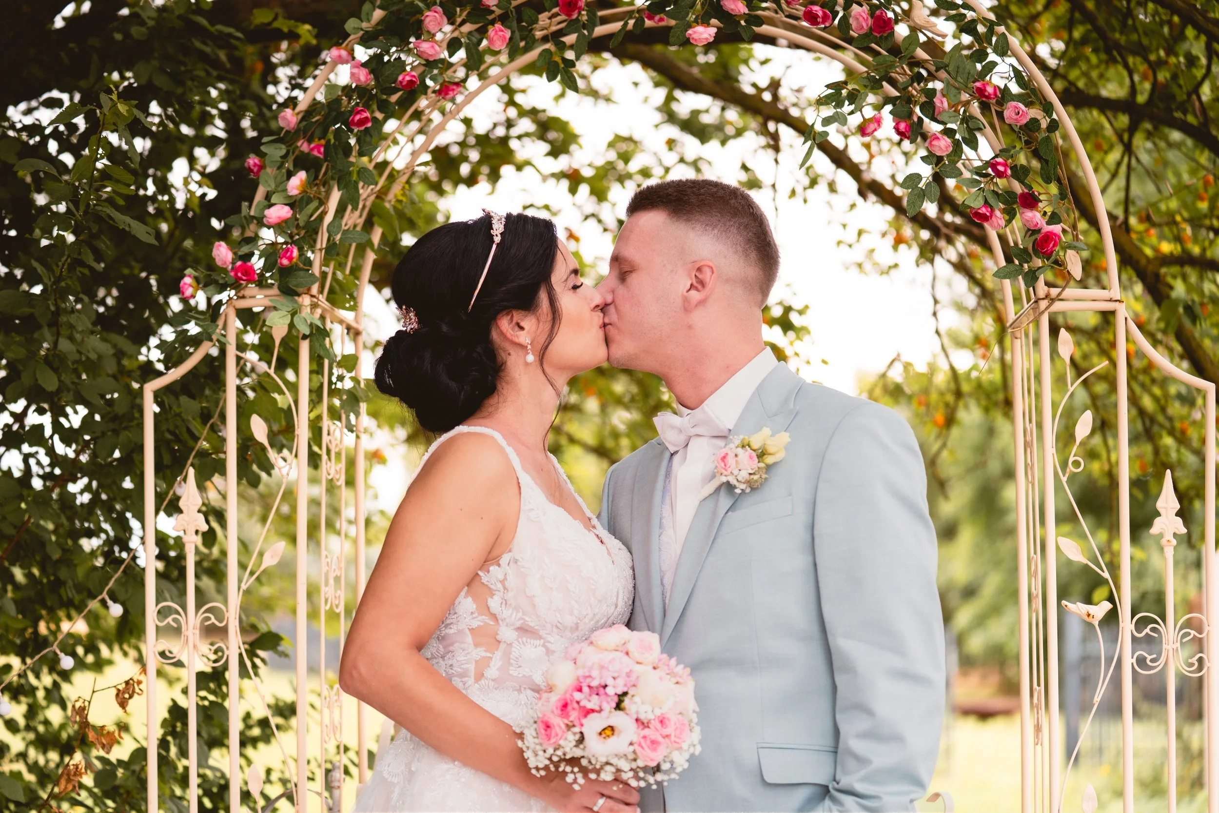 Ein frisch vermähltes Paar küsst sich vor einem blühenden Rosenbogen in einem Garten. Die Braut trägt ein weißes Kleid und eine Tiara, der Bräutigam einen hellblauen Anzug mit Fliege.