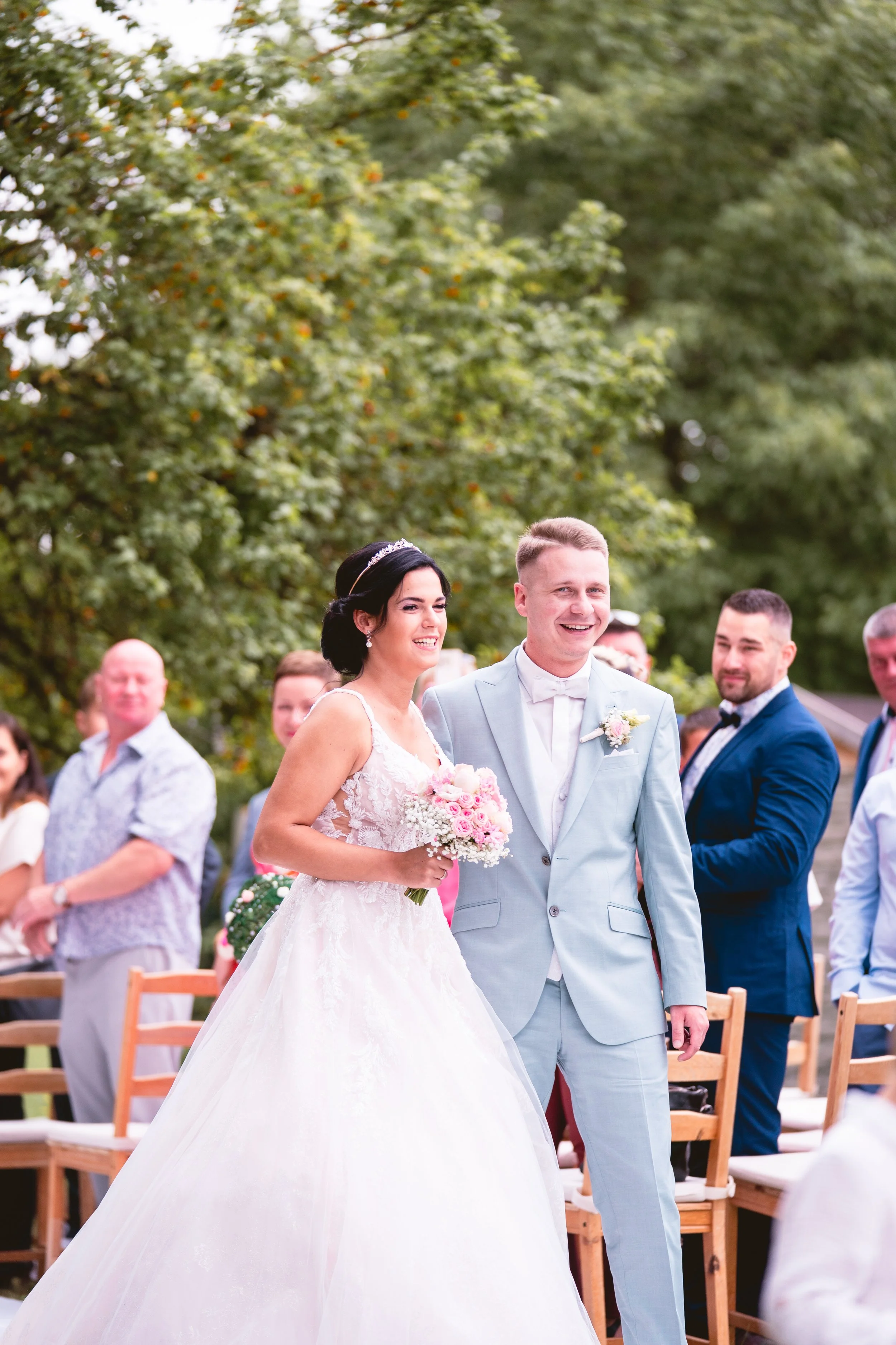Hochzeitspaar, bestehend aus einer Braut in weißem Hochzeitskleid mit Blumenstrauß und einem Bräutigam in hellblauem Anzug, umgeben von Hochzeitsgästen im Freien bei sonnigem Wetter, im Hintergrund grüne Bäume.