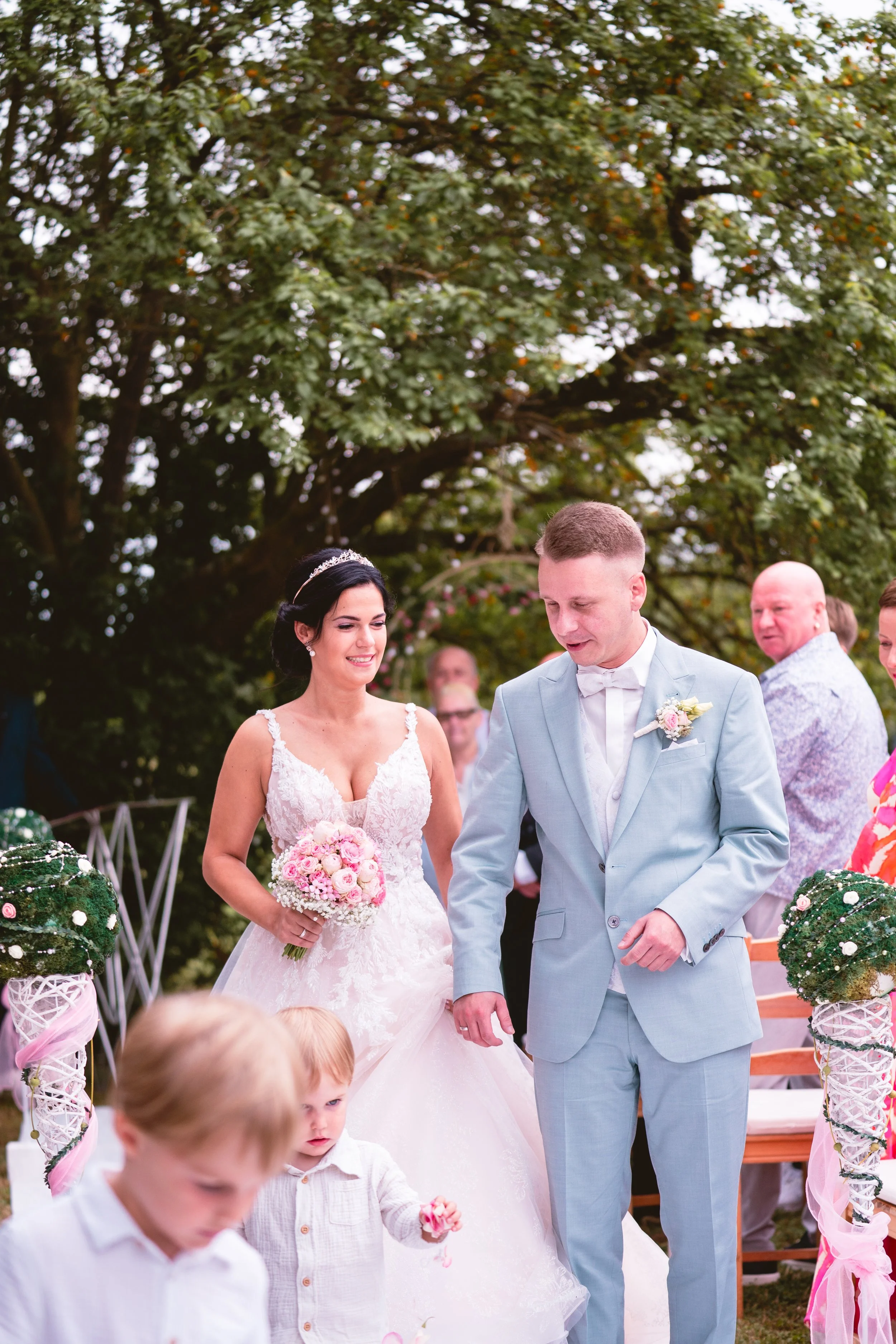 Ein Brautpaar bei einer Hochzeit im Freien, die Braut trägt ein weißes Hochzeitskleid mit Blumen und eine Tiara, der Bräutigam trägt einen hellblauen Anzug mit Fliege, im Hintergrund stehen Gäste und Bäume.