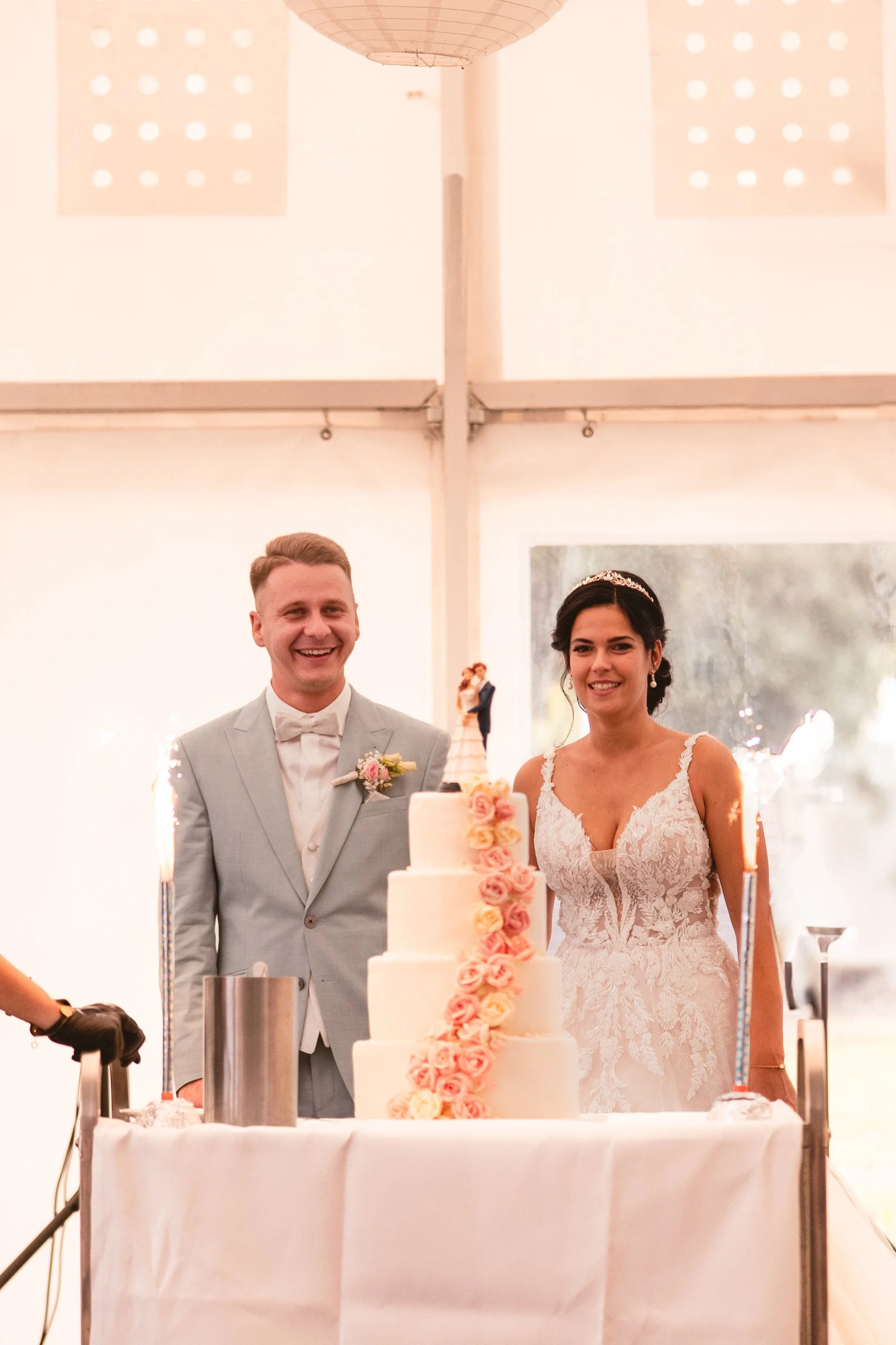 Ein Brautpaar bei ihrer Hochzeit, hinter einem mehrstöckigen Hochzeitstorte, im Hintergrund ein weißer Raum mit großen Fenstern, die Luftballons und Feuerwerkskörper.