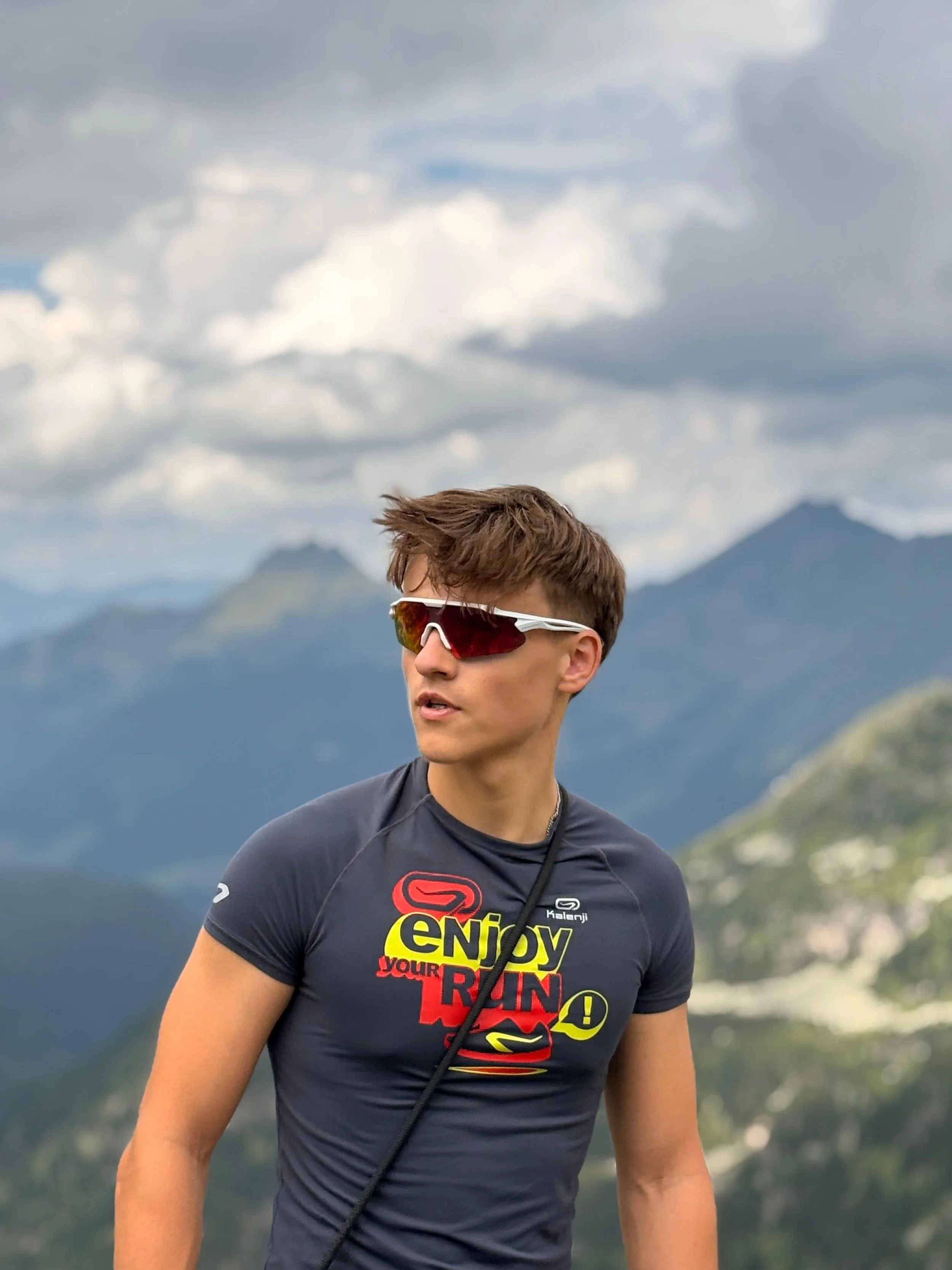 Ein junger Mann trägt eine schwarze Laufjacke mit buntem Aufdruck, Sonnenbrille und Kopfhörer, im Hintergrund Berge und Wolken, bei bewölktem Himmel.