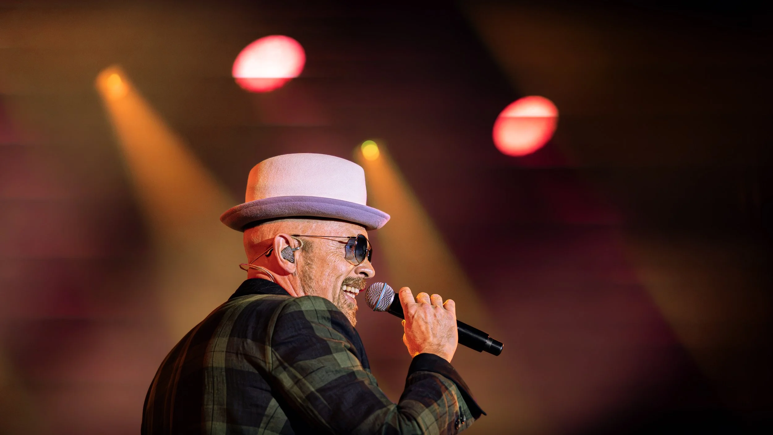 Ein Sänger mit Hut und Sonnenbrille singt in einem Konzert, im Hintergrund sind bunte Bühnenlichter.
