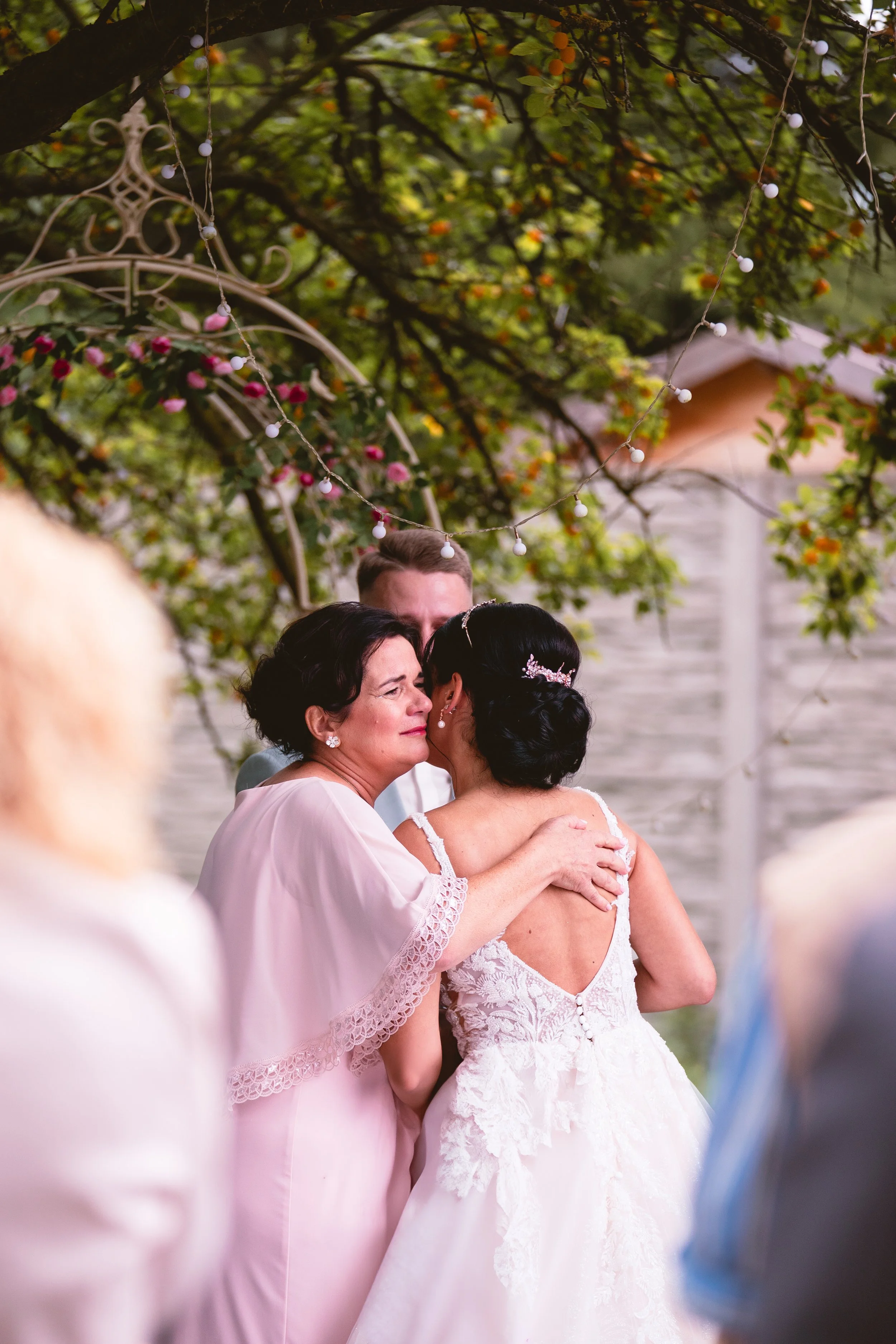 Eine emotionale Szene bei einer Hochzeit, in der eine ältere Frau die Braut zärtlich umarmt, während sie unter einem Baum mit Lichterketten steht. Im Hintergrund sind weitere Gäste zu sehen.