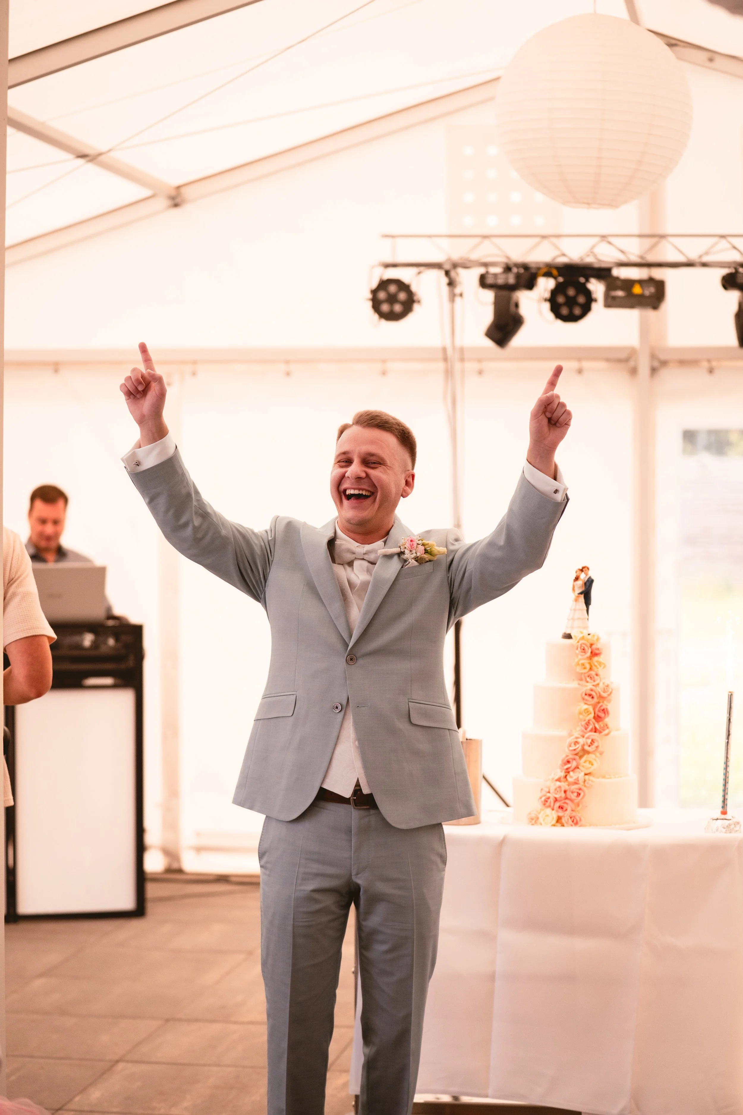 Ein lachender Mann in einem grauen Anzug mit einem Blumenanstecker am Revers hebt die Arme hoch, wahrscheinlich bei einer Feier, neben einem mehrstöckigen Hochzeitstorte mit Rosen Deko, im Hintergrund sind DJ und DJ-Equipment sichtbar.