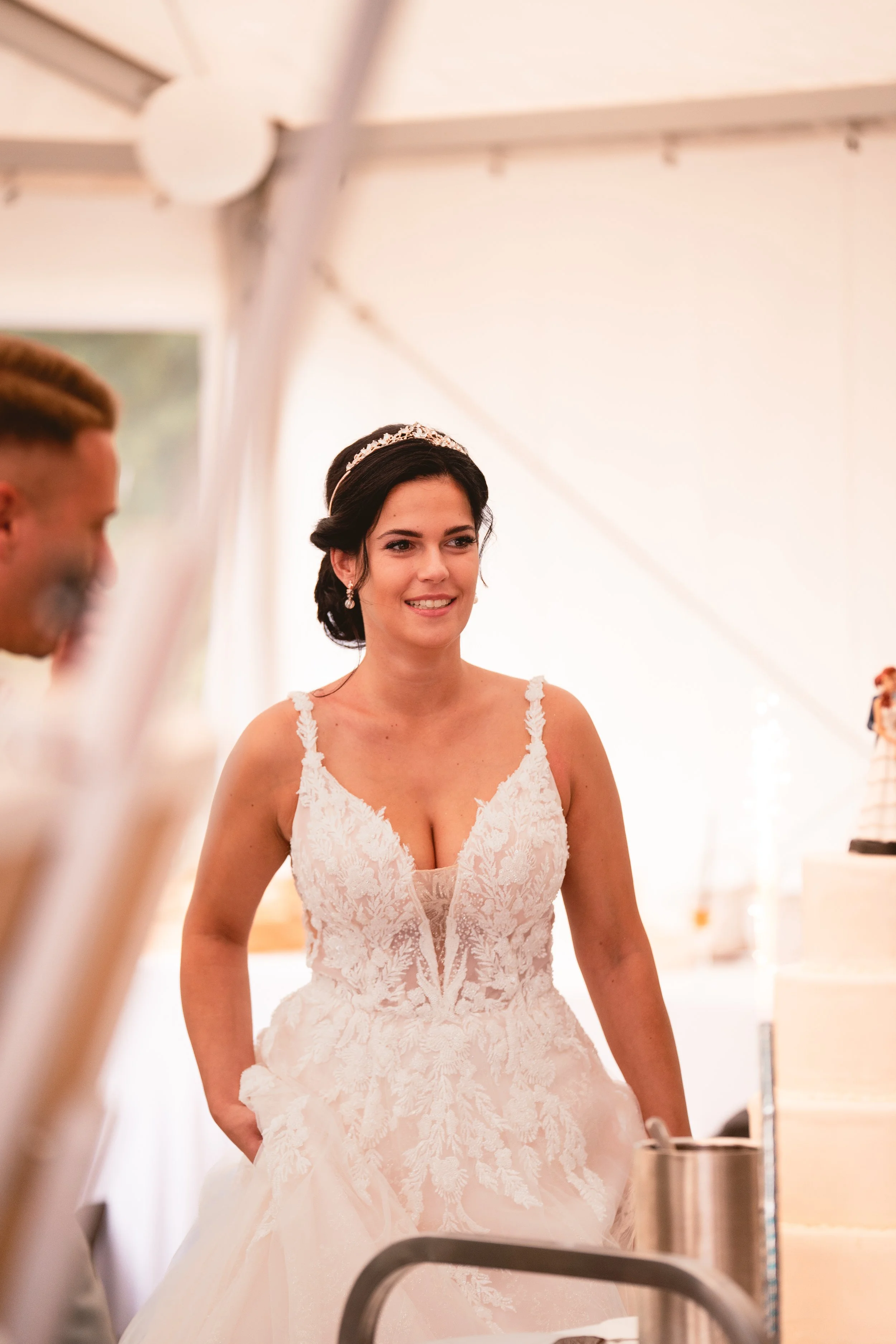 Eine Frau trägt ein weißes Hochzeitskleid mit floralen Mustern, eine Tiara und Ohrringe, während sie in einer Zelt- oder Festhalle steht.