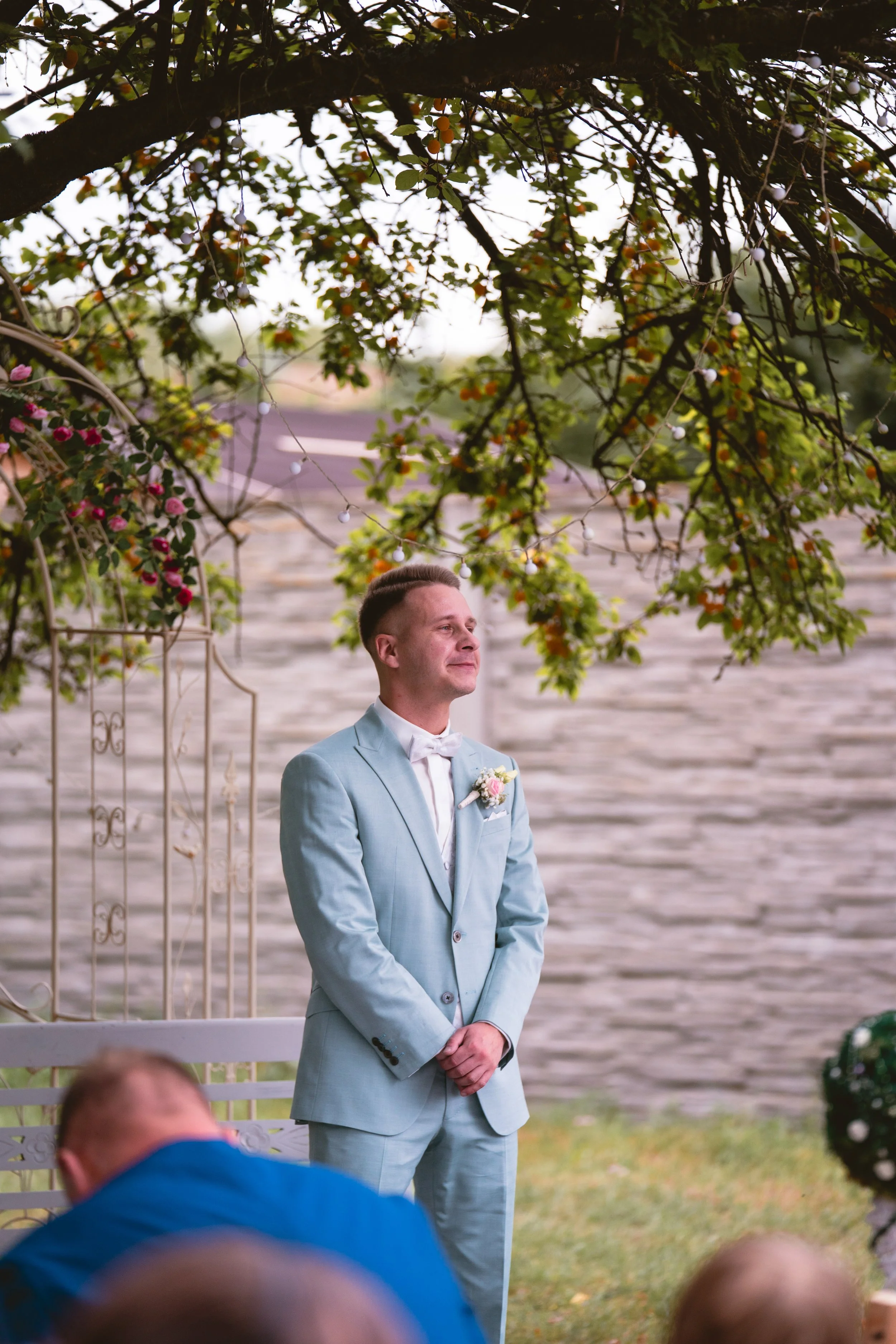 Ein Mann in einem hellblauen Anzug mit Fliege, der bei einer Hochzeit oder formellen Veranstaltung steht, im Hintergrund sind Bäume mit Lichterketten, eine weiße Sitzbank und eine Mauer.