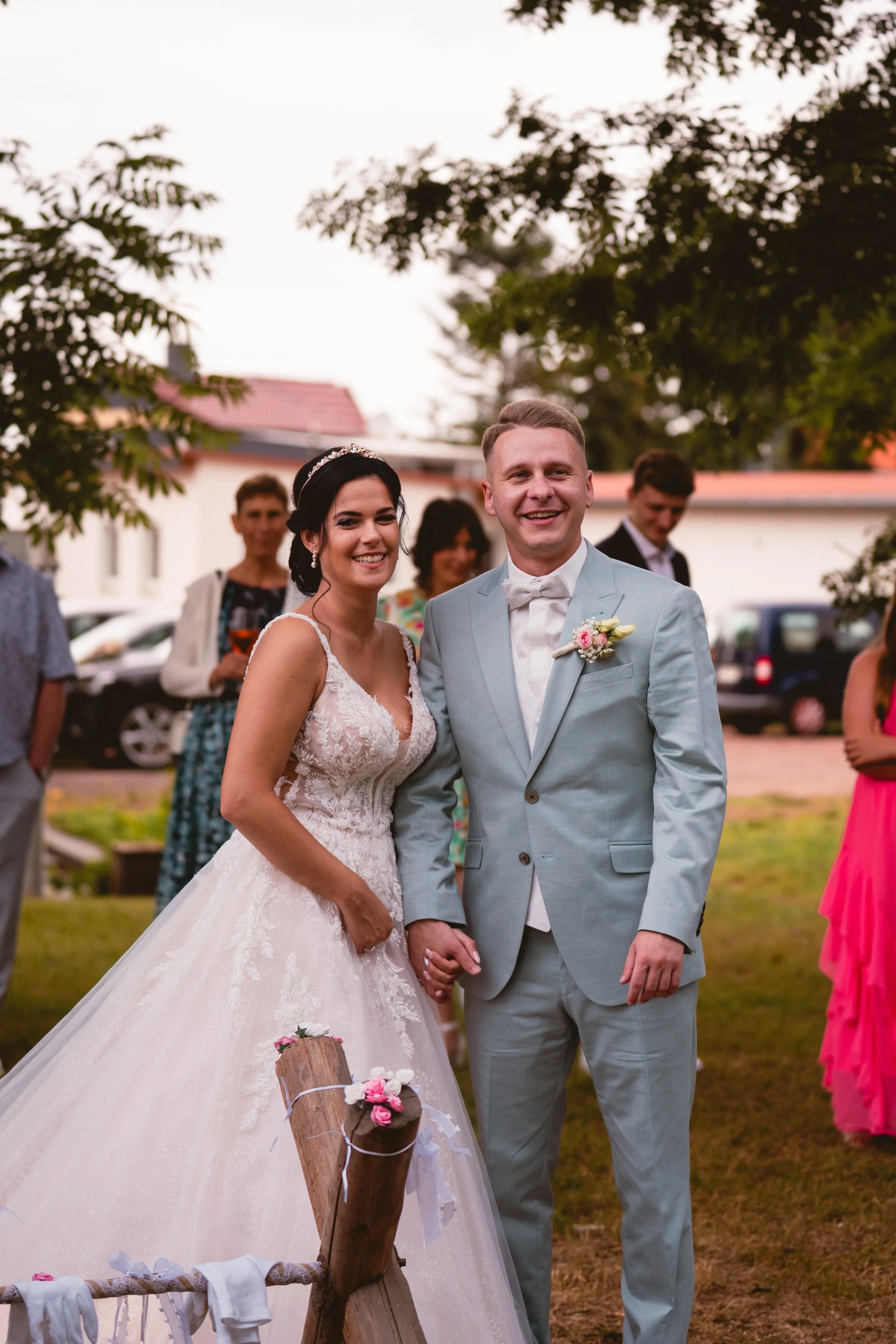 Braut und Bräutigam stehen bei einer Hochzeit im Freien, umgeben von Gästen, die im Hintergrund stehen. Sie halten sich an den Händen, lächeln glücklich und tragen formelle Kleidung, die Braut trägt ein weißes Brautkleid mit Spitze und eine Tiara, de