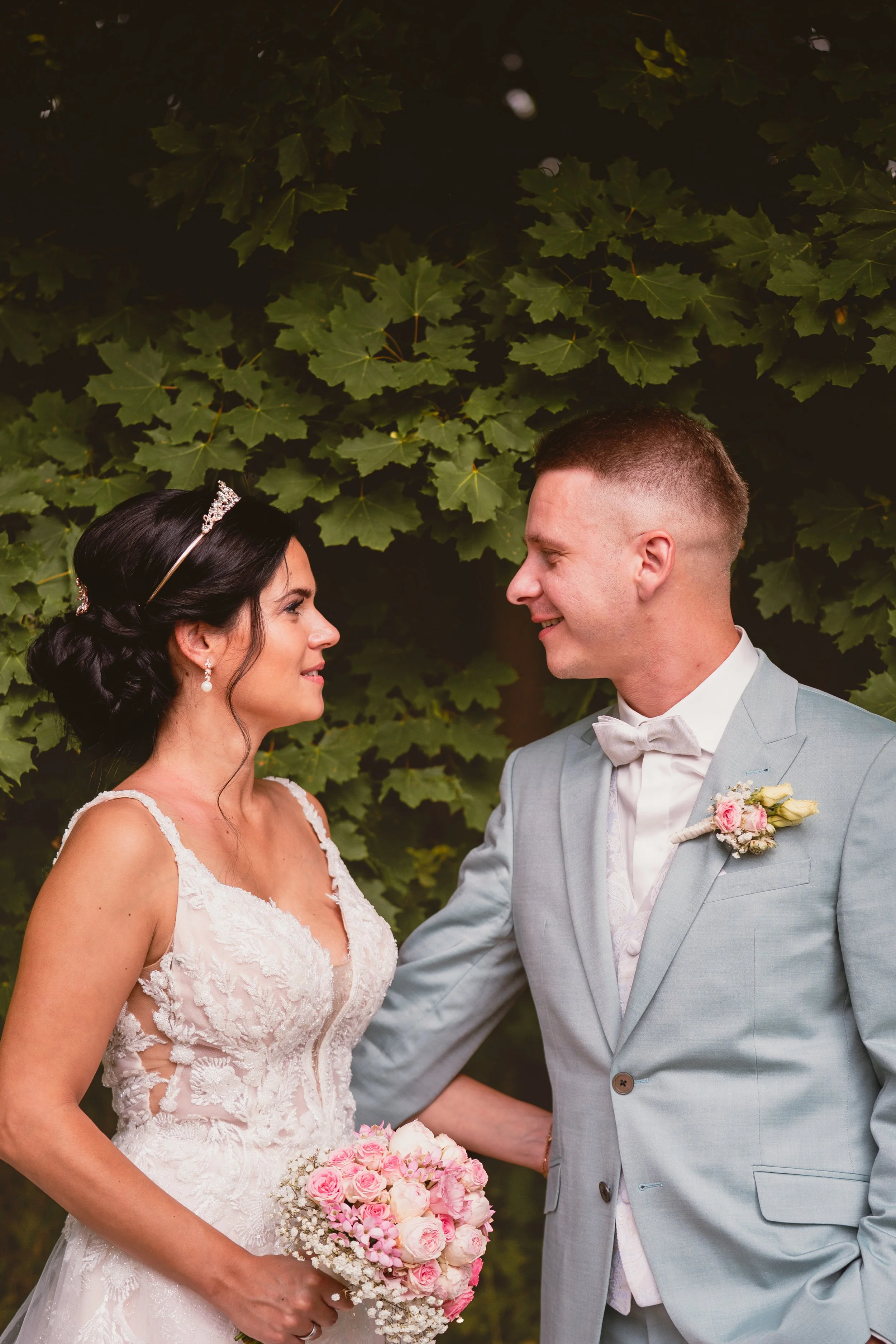 Ein Brautpaar steht sich gegenüber, die Braut trägt ein weißes Hochzeitskleid mit Spitze und hat dunkle Haare, die zu einem Zopf gestylt sind. Sie trägt einen Tiara und Ohrringe. Der Bräutigam trägt einen hellgrauen Anzug mit weißem Hemd, Fliege und 