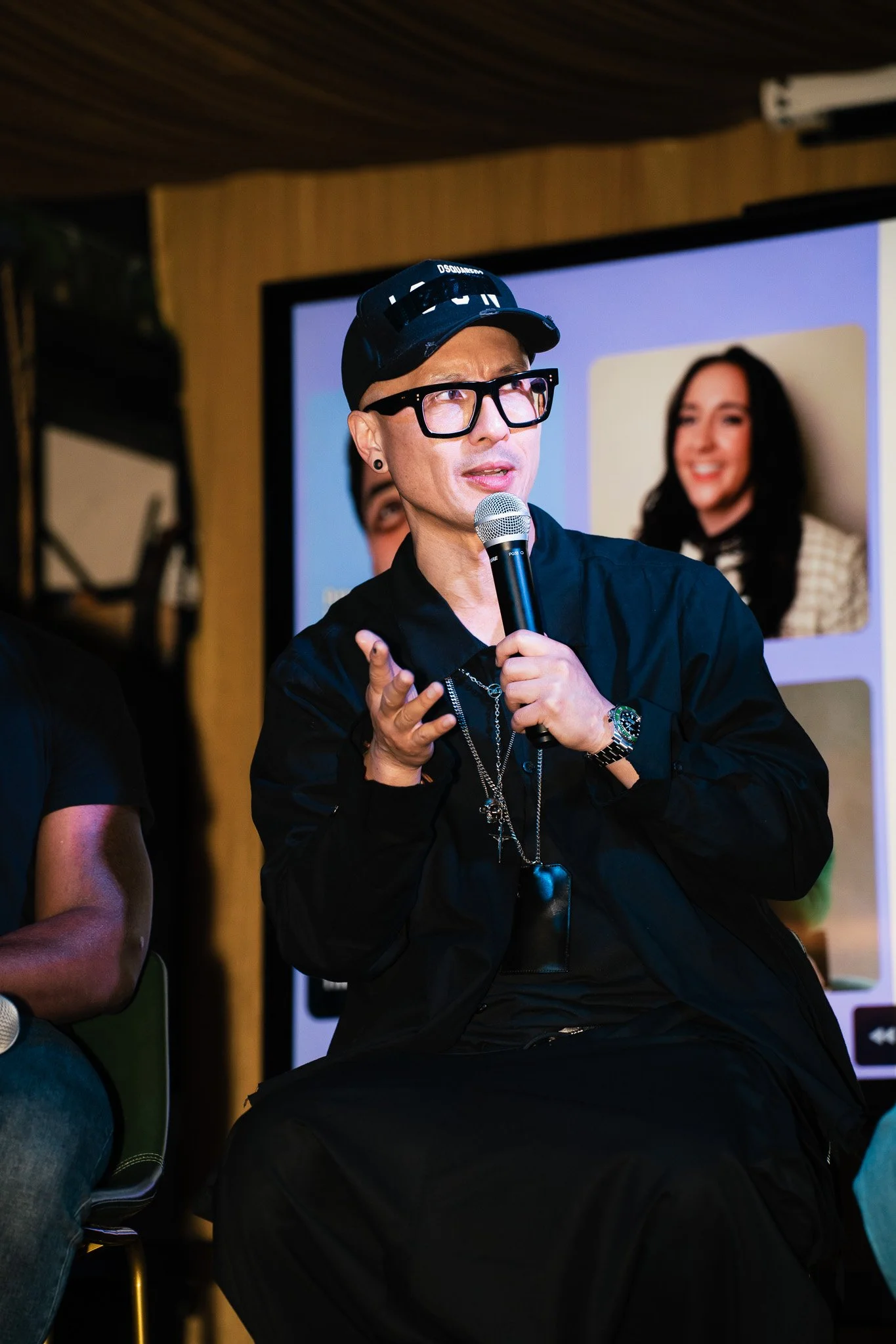 A man with glasses, earrings, and a black hat with white text, holding a microphone and speaking at an indoor event. A large screen behind him displays a woman smiling.