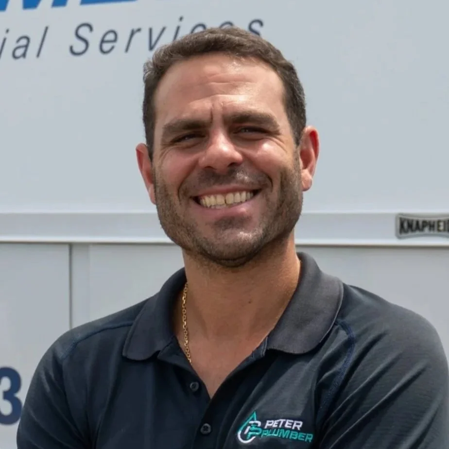 A smiling man with dark hair, wearing a black collared shirt with a company logo, standing in front of a sign with partially visible text.