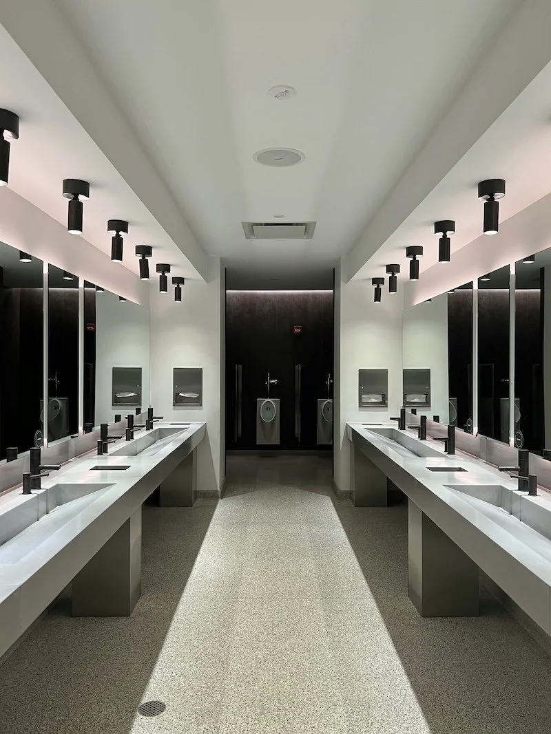 Modern public restroom with white sinks, black faucets, large mirrors, and hand dryers on the walls.