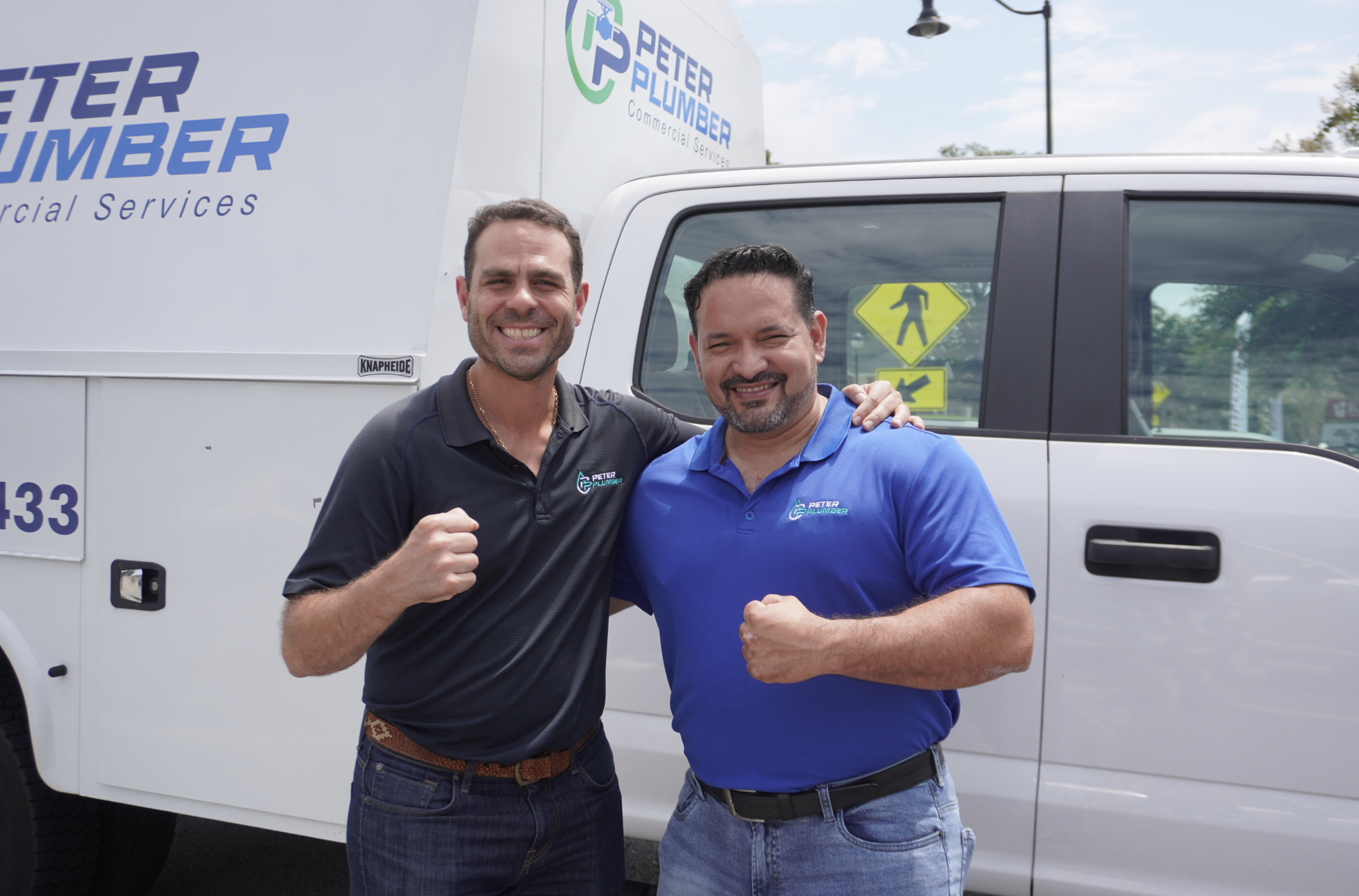 Two smiling men in Peter Plumber uniforms standing in front of a Peter Plumber service truck, posing with fists clenched in a celebratory gesture.