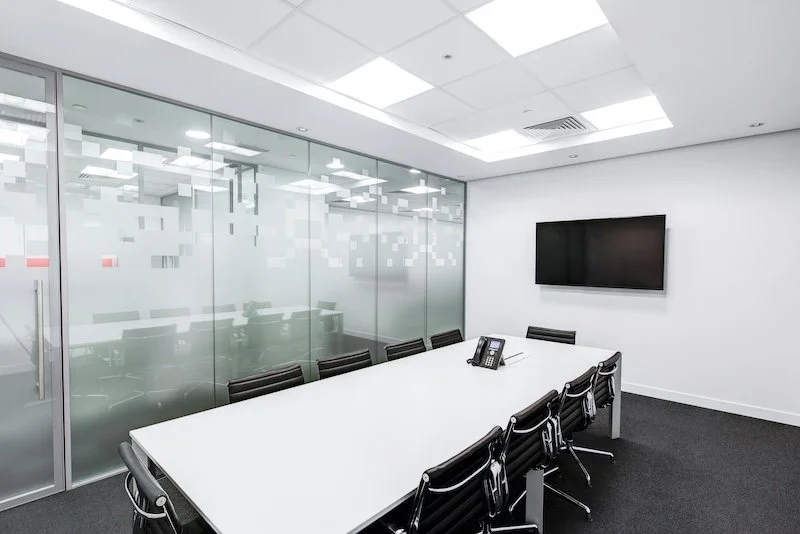 Empty modern conference room with white table, nine black chairs, glass wall, large flat-screen TV, and phone on table.