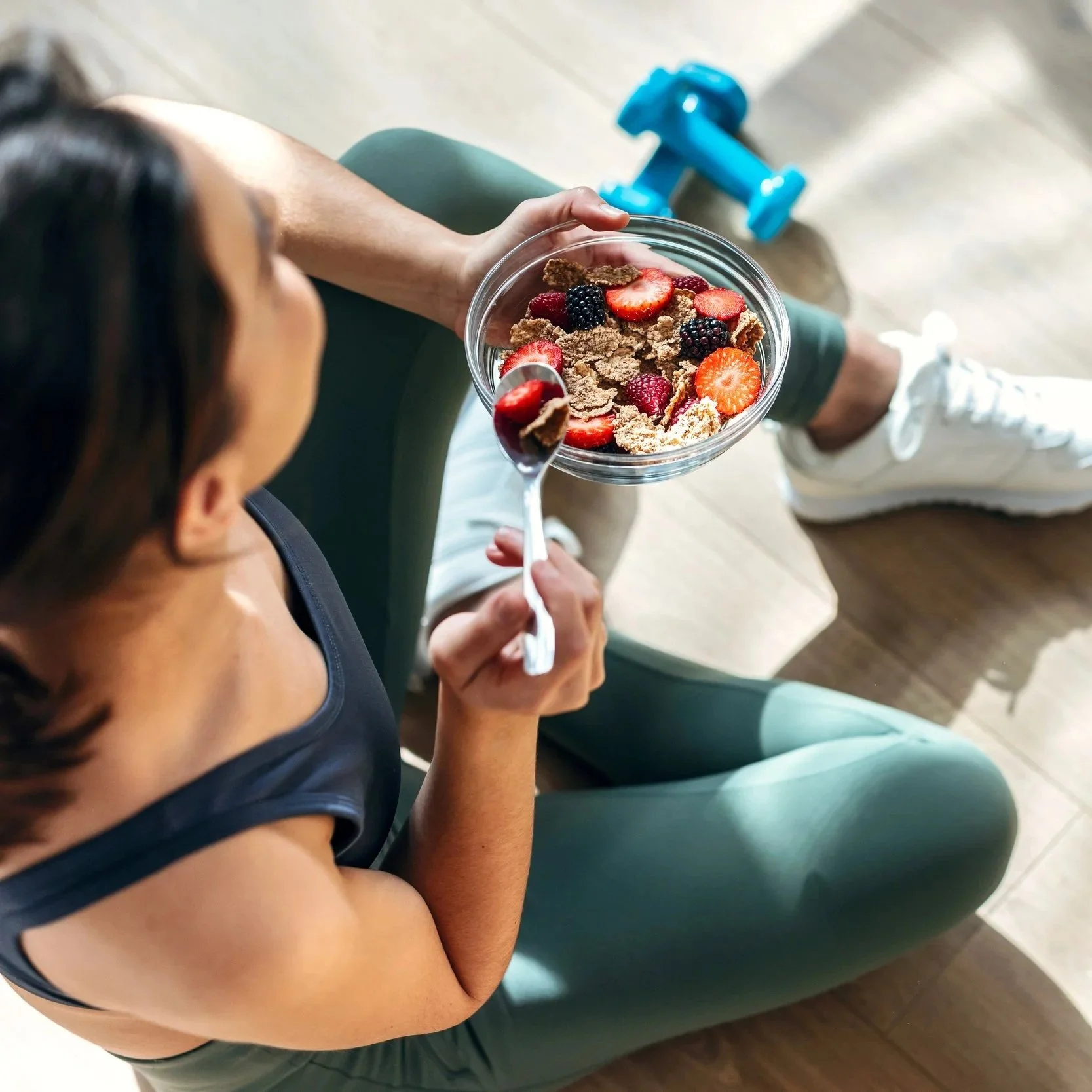 A woman sitting on a green exercise ball, holding a glass bowl of cereal with strawberries, blackberries, and raspberries, and a spoonful of cereal and fruit.