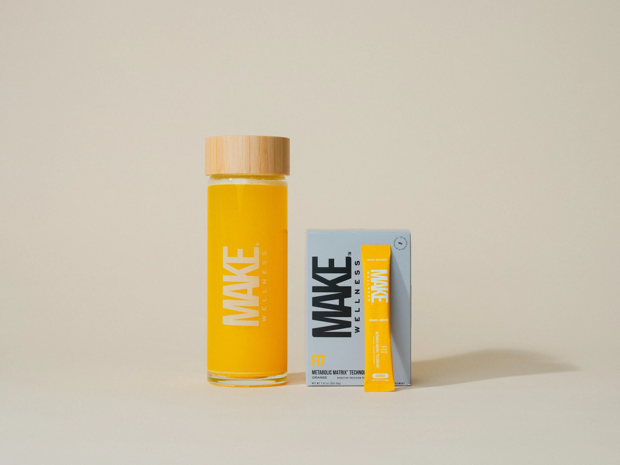 A bottle of MNAKE Wellness supplement and a box of MNAKE supplements with an orange flavored stick in front of the box, on a minimalist light-colored background.