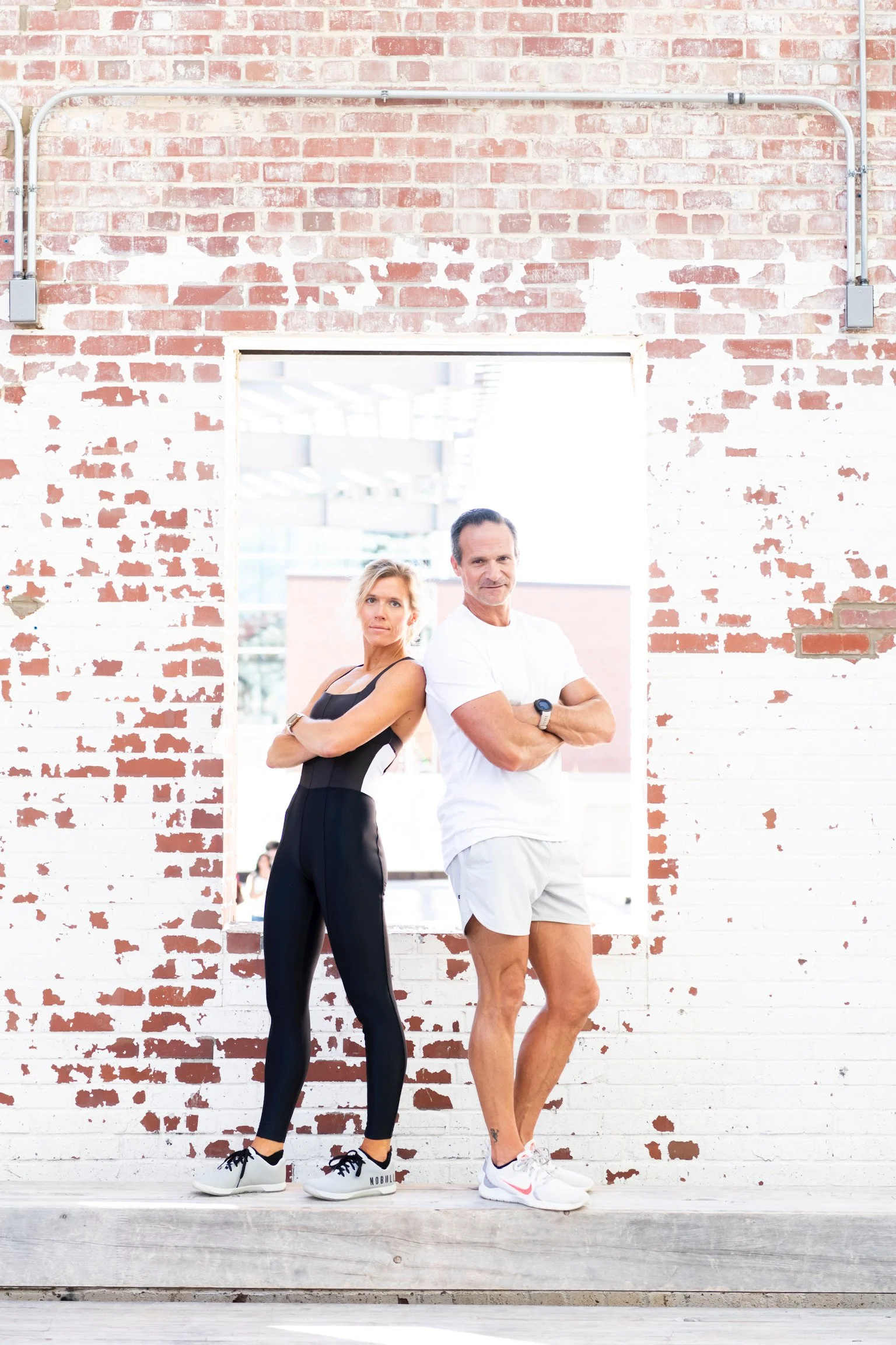 A woman and a man standing back-to-back with crossed arms in front of a brick wall, inside a bright gym or studio.