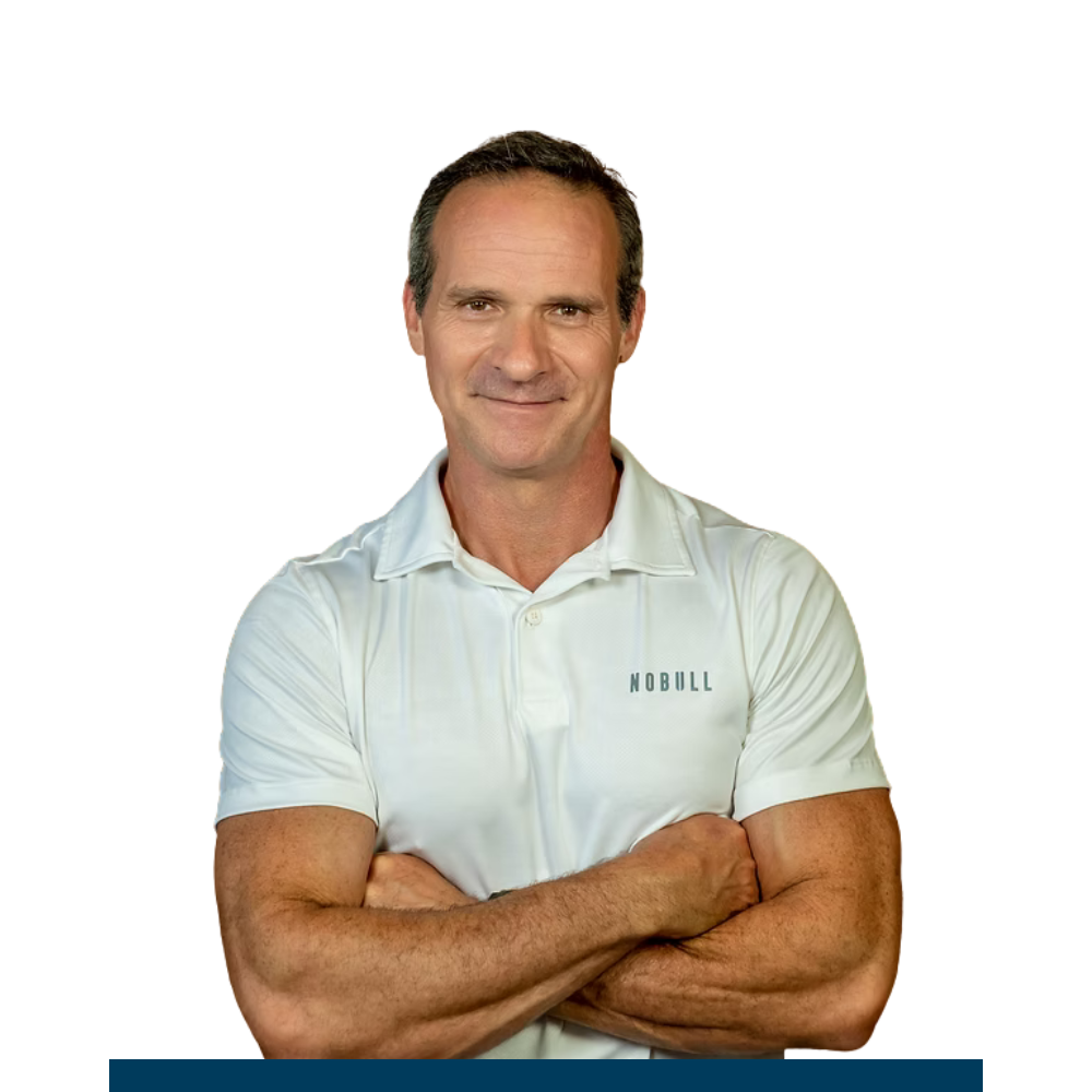 A middle-aged man with short dark hair, smiling, wearing a white short-sleeved polo shirt with the word NOBULL on it, crossing his arms.