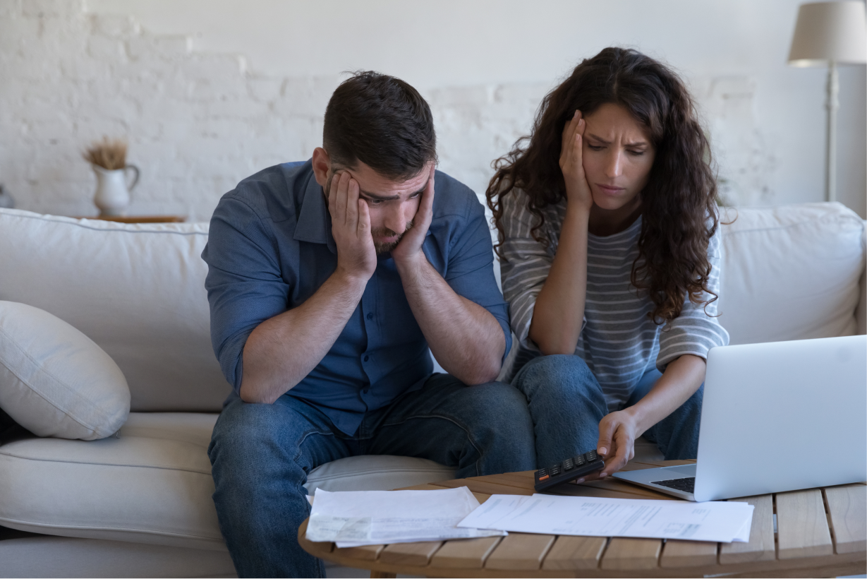 Stressed couple sitting on couch reviewing bills with laptop and paperwork