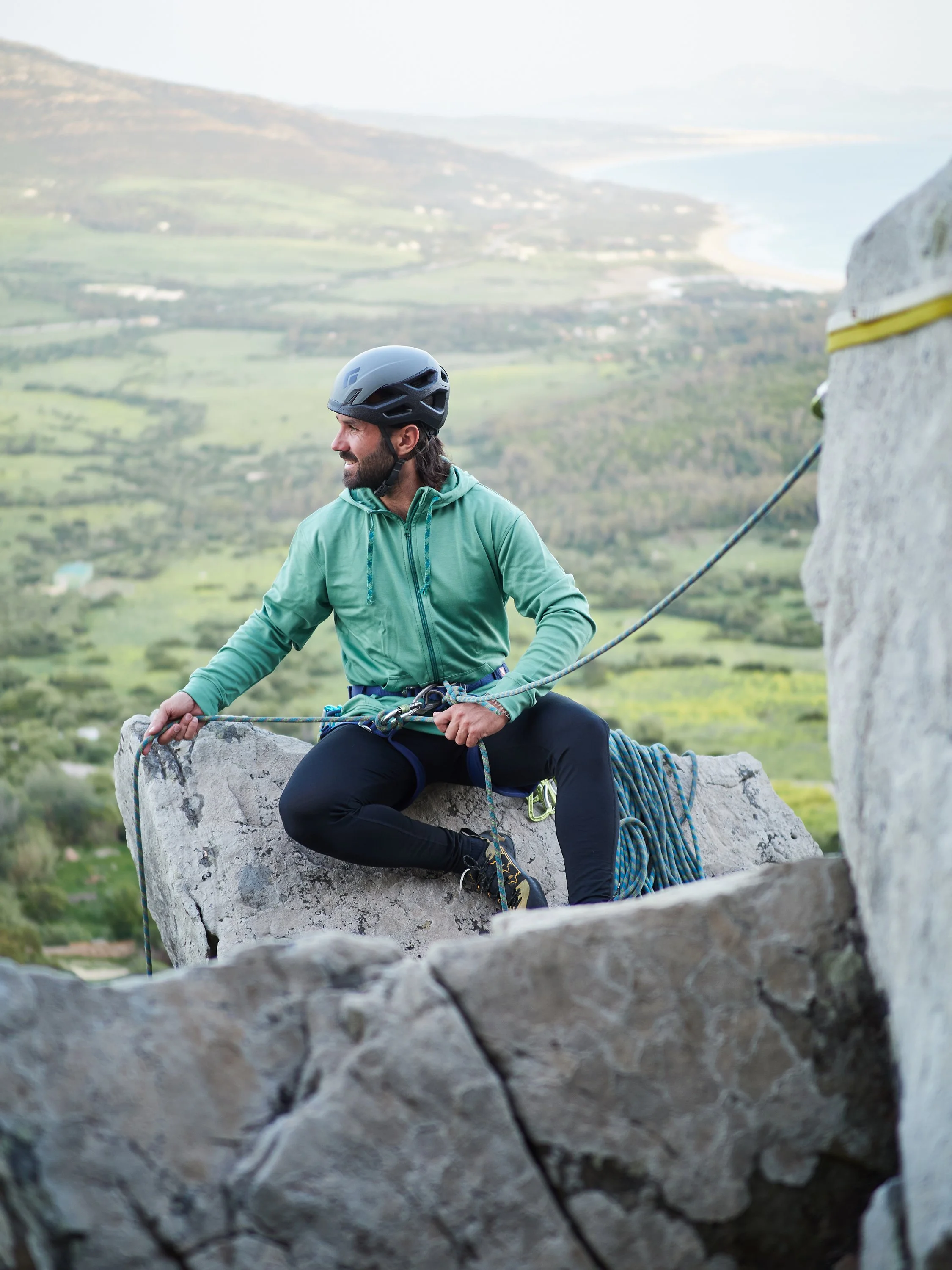 Kletterer mit Helm und Kletterausrüstung auf einem Felsen mit Blick auf eine grüne Landschaft und Wasser im Hintergrund.