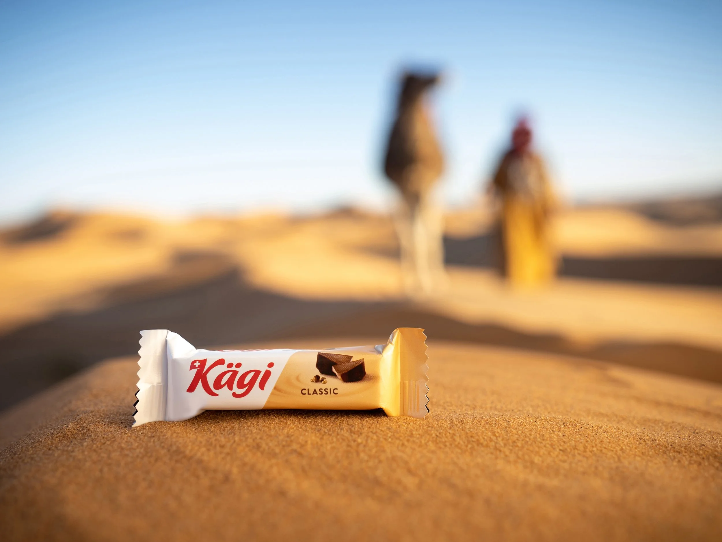 Ein Kagi Classic Schokoladenriegel liegt auf Sand in der Wüste, im Hintergrund sind zwei Menschen in traditioneller Kleidung mit Kamelen. Der Himmel ist klar und blau.