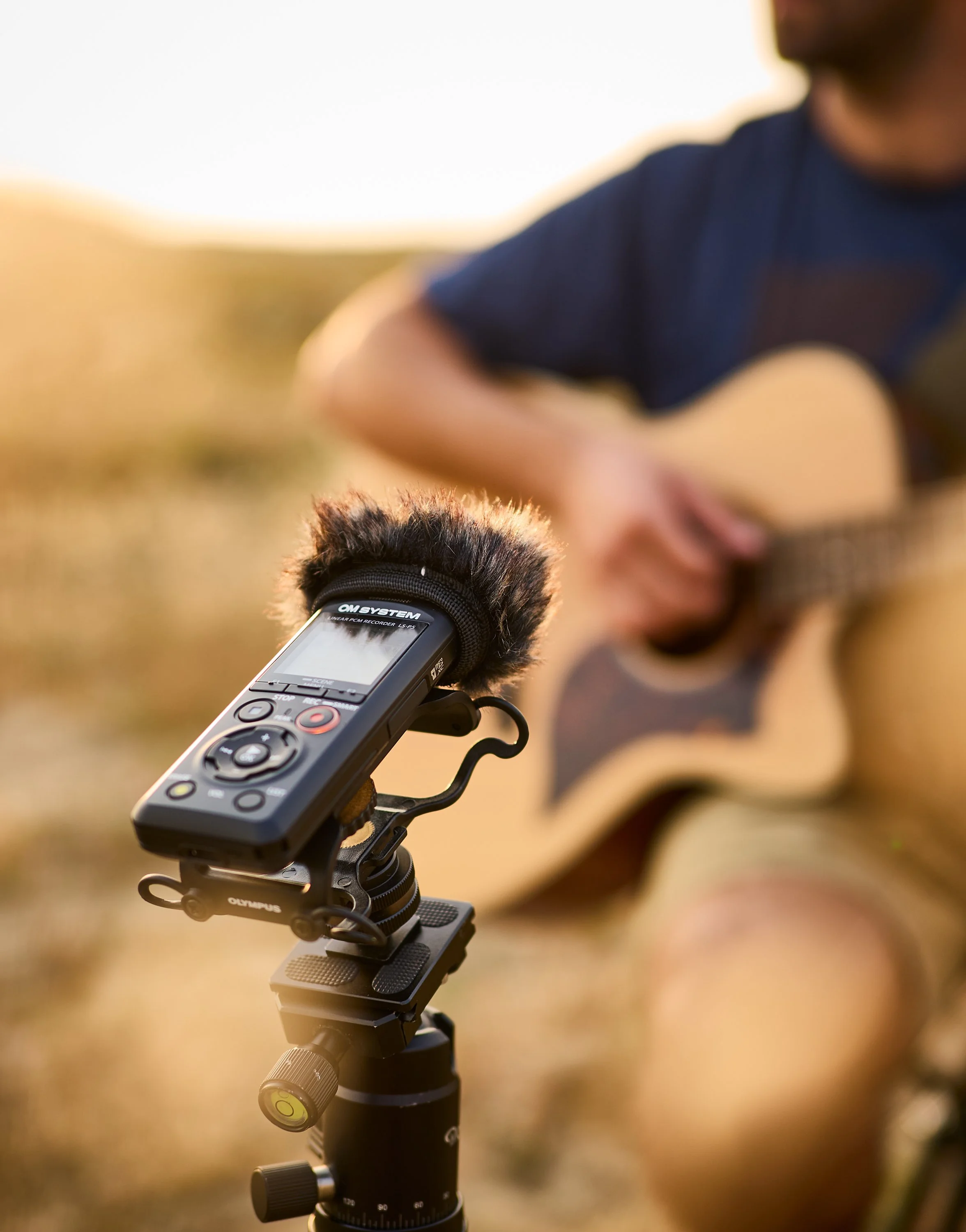 Nahaufnahme eines Audio-Recorders auf einem Stativ, im Hintergrund ein Musiker, der Gitarre spielt, bei Sonnenuntergang im Freien.