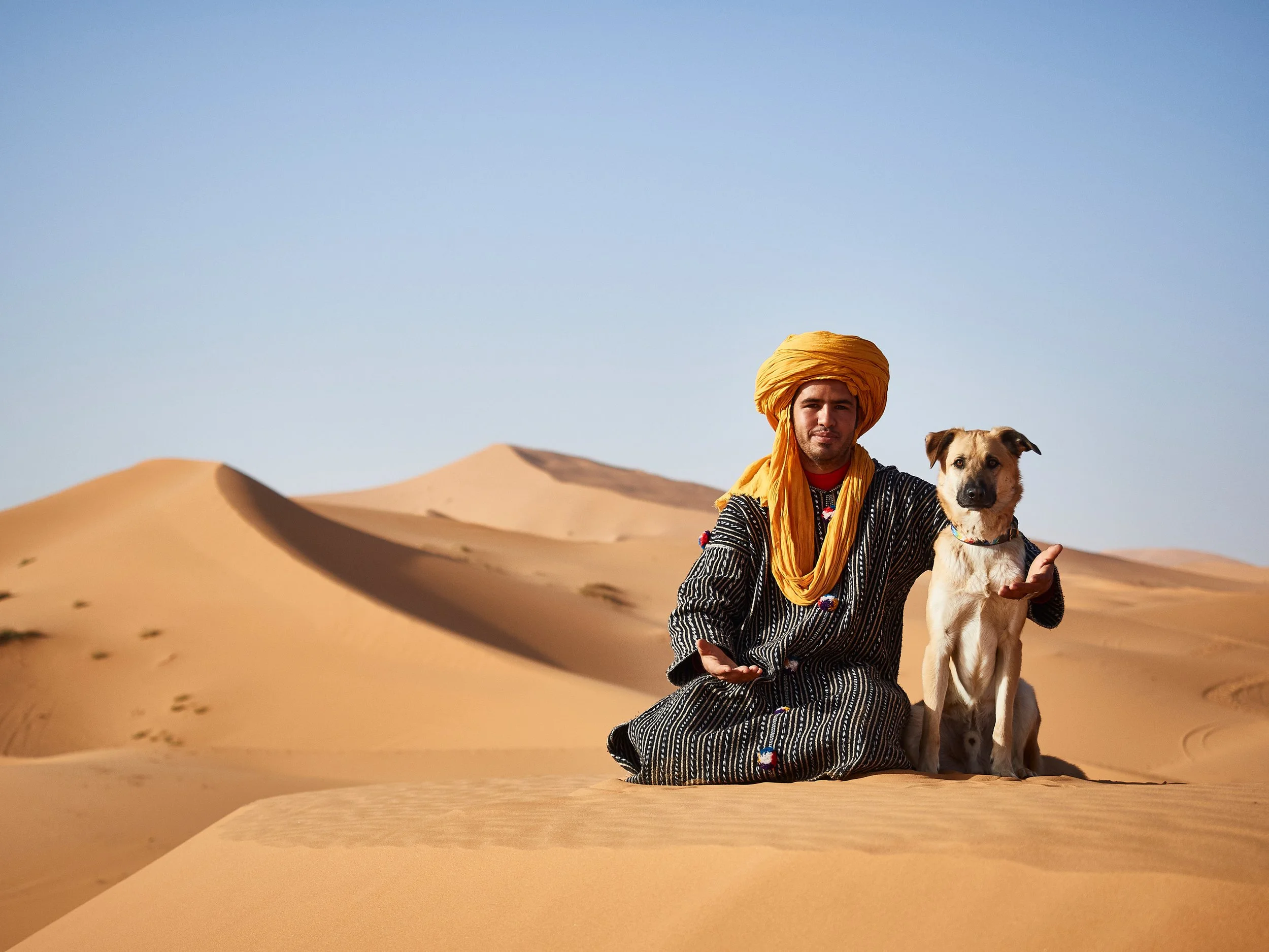 Mann in traditioneller Kleidung mit gelbem Turban im Sanddünen-Wüste, neben einem Hund sitzend.