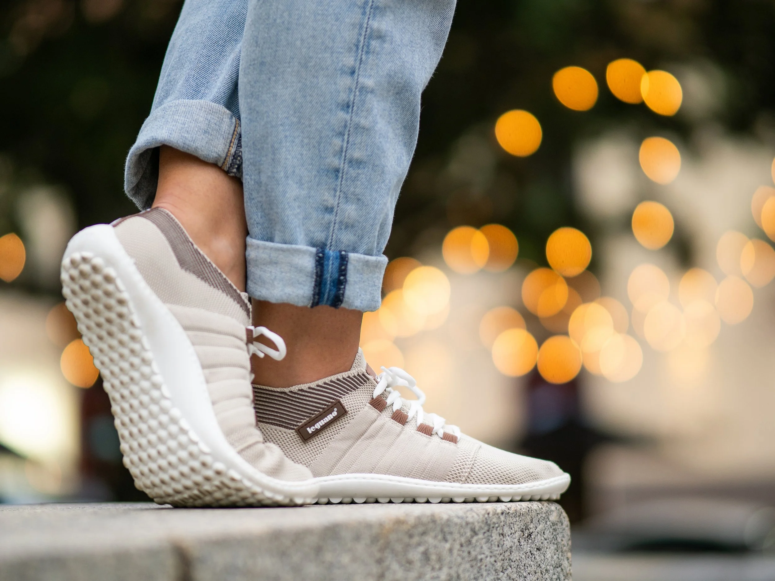 Nahaufnahme von weißen Sportschuhen mit braunen Akzenten und Jeanshose, die an einer Stadtbrüstung stehen, mit unscharfen Lichter im Hintergrund.